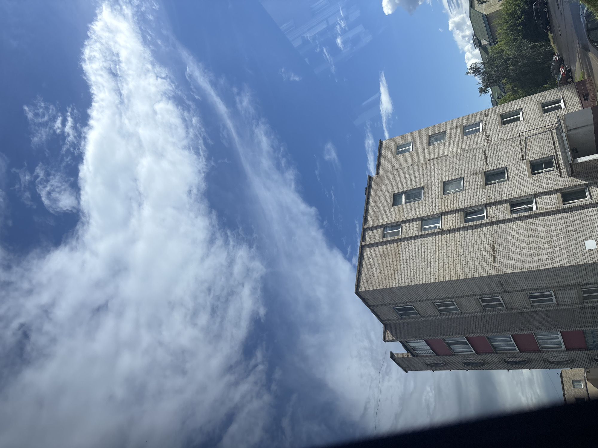 The image depicts a view from an elevated position, looking upwards and outwards towards a brick apartment building against a bright blue sky filled with dynamic white clouds. The apartment building is light gray brick with some reddish-brown sections. It has multiple windows, most of which appear to be closed and reflect the sky. The building's facade shows signs of wear, with some visible cracks and discoloration. To the left of the main building, another smaller building with a white facade and a single window is partially visible.
In the lower right quadrant of the image, a street scene is visible, with parked cars, including a dark SUV and a red sedan. Green trees and other buildings with green roofs are present in the background, suggesting a residential urban or suburban setting. The lighting indicates it is daytime, with bright sunlight casting shadows. The image appears to have been taken through a window, as evidenced by a dark, blurred area on the left and a subtle reflection on the right. This is a scene in the city of Kapyĺ, Belarus.