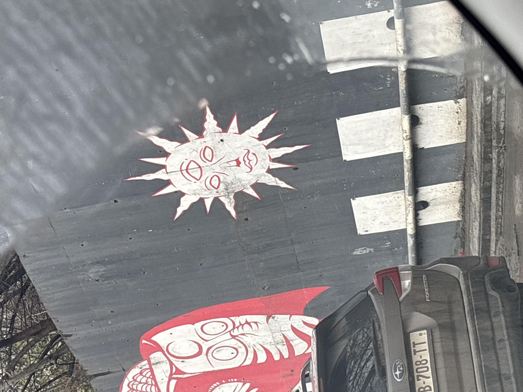 The image displays an outdoor urban scene in Tbilisi, Georgia, captured through a vehicle window. A dark grey concrete retaining wall, showing signs of wear and staining, forms the backdrop. Two distinct pieces of street art are visible on this wall. On the left, a large, stylized red and white face or mask, featuring prominent circular eyes, is partially depicted. To its right, a white sun-like figure with radiating points, outlined in red, showcases a facial representation with three eyes, a nose, and a small mouth. Below the wall, a roadside barrier composed of vertical panels with alternating black and white stripes and a horizontal metal rail is present. In the lower-left quadrant, the rear-left section of a dirty, dark grey Subaru Forester SUV is visible, displaying a Georgian license plate "B-708-TT". Above the wall, in the upper-left, several bare tree branches extend into the frame. The foreground exhibits reflections and distortions consistent with a car windshield or window, and the ground surface appears damp.