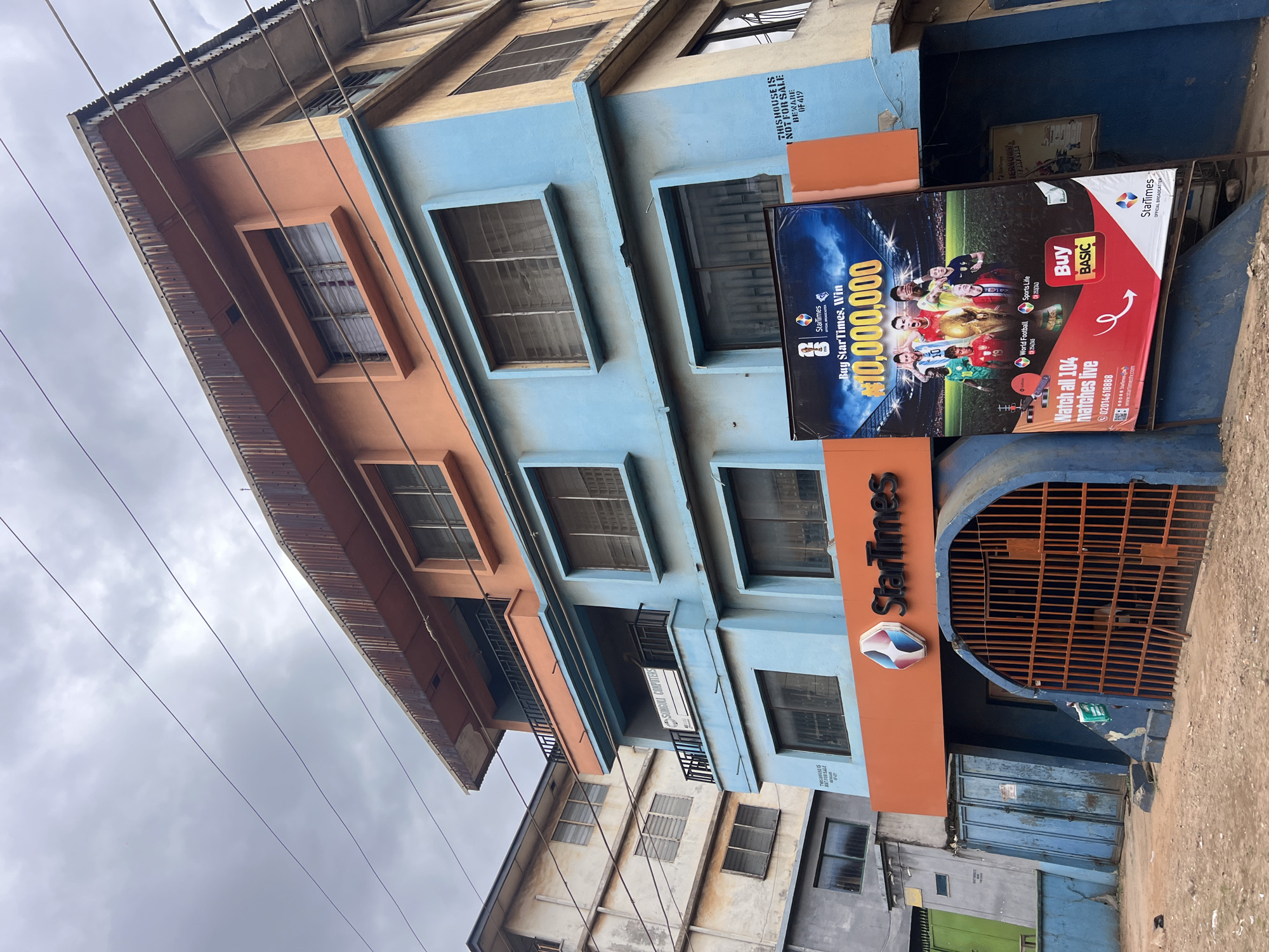 This is a full, eye-level, outdoor shot of the StarTimes office in Nkpor, Nigeria, on a cloudy day. The building has three visible floors, with the lower two painted in a pale blue and the top floor in a pale orange. The exterior of the building is weathered, with some peeling paint and rust evident on the corrugated iron roof overhangs. A large banner advertising StarTimes and a football match promotion is prominently displayed on the lower front of the building. The banner features images of famous football players and a large sum of money, promoting a contest. Below the banner, a blue, arched entranceway with orange bars is visible, leading into the building. To the left of the entrance are blue metal doors. The ground in front of the building is dirt and gravel. In the background, other buildings can be seen, some appearing to be in a state of disrepair. There are no people visible in the frame. Overhead power lines crisscross the sky.
