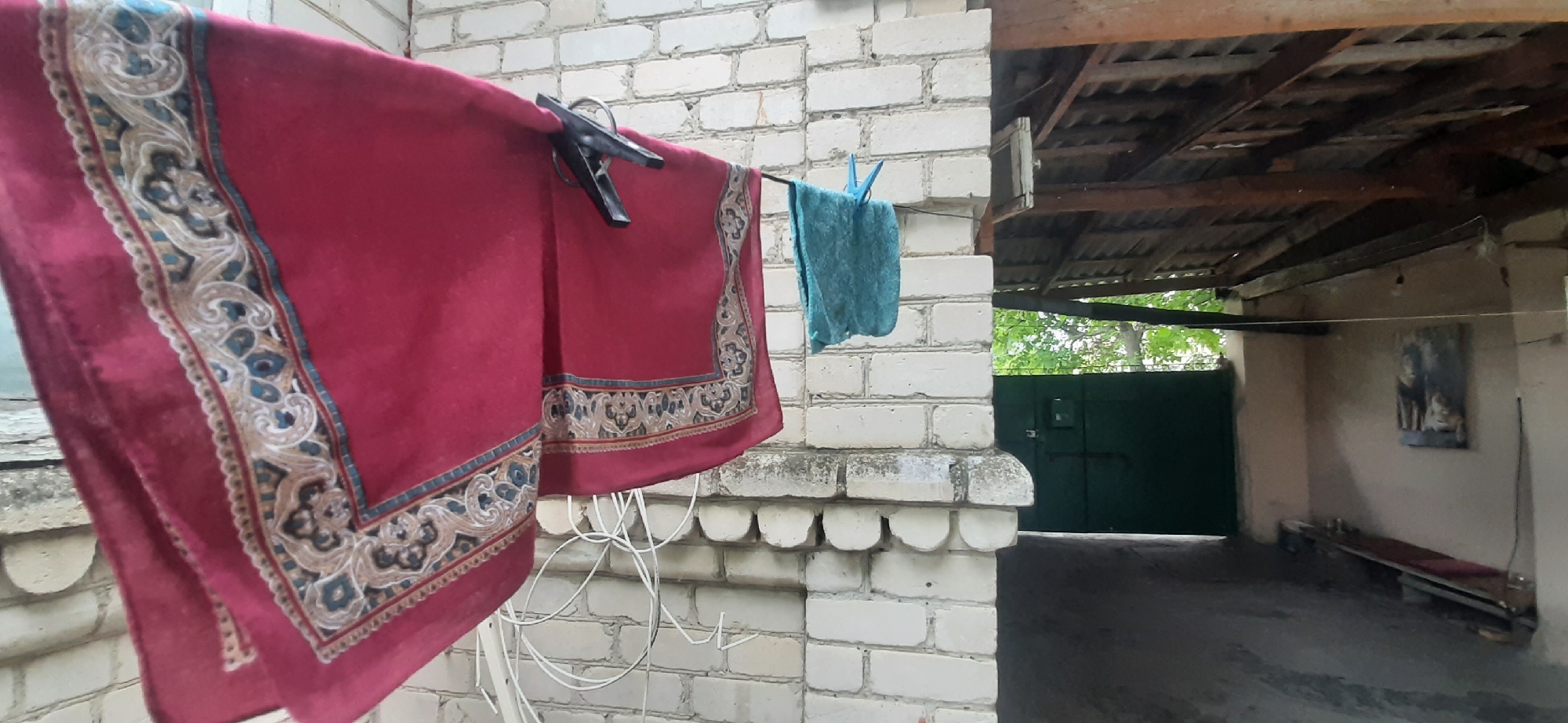 A red and gold patterned cloth and a teal towel are hanging on a clothesline attached to a white brick wall. A black clothespin is on the red cloth, and a blue clothespin is on the teal towel. To the right of the wall, a dark green metal gate is visible behind a wooden roof structure. A long wooden bench is against a wall in the background, with a framed picture of a lion and cub hanging above it. The scene is likely a backyard or service area in Yessentukskaya, Russia.
