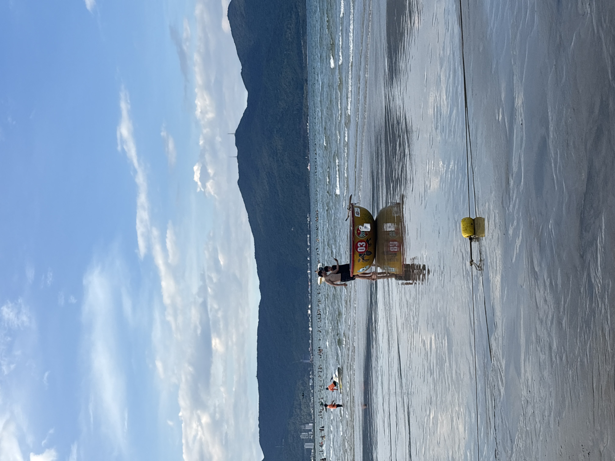 The image depicts a beach scene in Đà Nẵng, Vietnam. In the foreground, a circular, yellow and red basket boat with the number "03" is partially submerged in shallow water. A rope with a yellow float is visible extending across the wet sand. Two men, one appearing to be holding a phone, stand beside the boat. In the middle ground, the sea is choppy with moderate waves. Further out, a crowd of people are visible in the water and on the beach, with some engaging in water activities like riding a jet ski and standing in the surf. In the background, a lush, green mountain range with some buildings and radio towers on its slopes forms the horizon. The sky is partly cloudy with blue patches.
