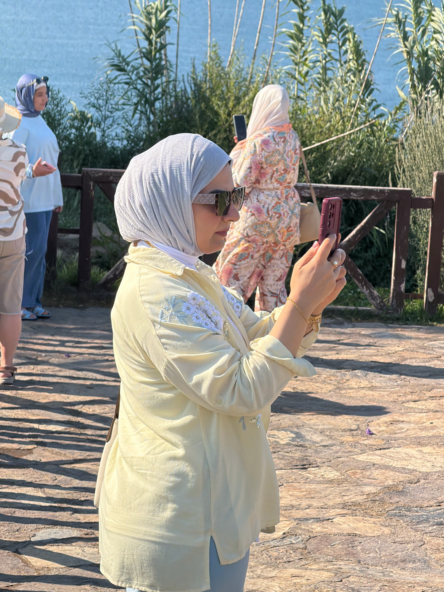 In Antalya, Turkey, on a sunny day, a woman wearing a light blue hijab and a light blue long-sleeved shirt is standing near a body of water. She is looking at something in her hand, possibly a phone. Behind her, another woman in a colorful, floral-patterned dress and a light colored hijab is holding a black smartphone. In the foreground, a woman wearing a light yellow, long-sleeved shirt and a light gray hijab is taking a photo with her maroon smartphone. She is wearing sunglasses and a bracelet on her left wrist. The ground is paved with stones. There are tall green plants and trees in the background. A wooden fence runs through the scene.