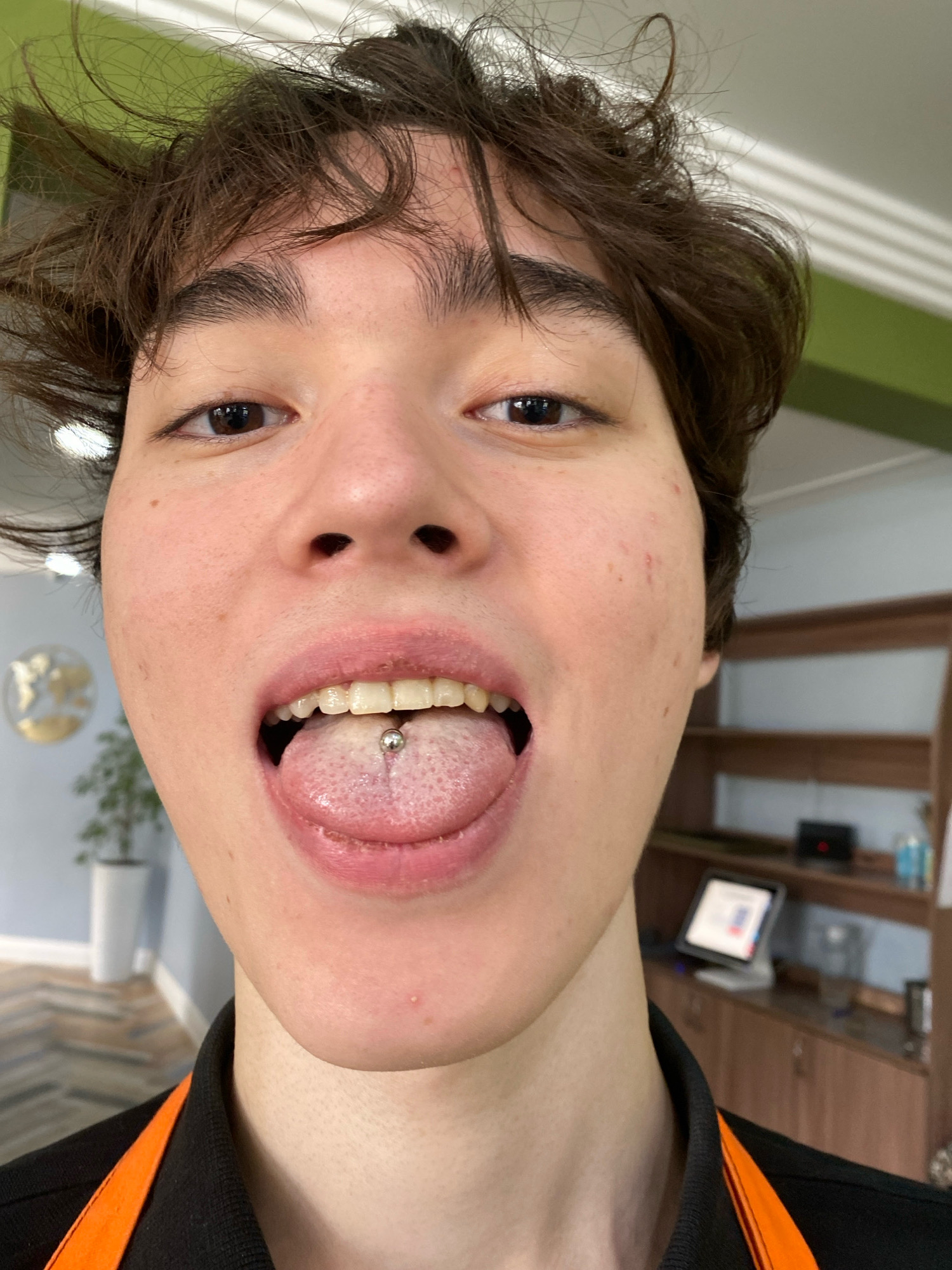 A close-up, eye-level, medium shot captures a young person with brown, tousled hair sticking out from the top of their head. Their mouth is open, revealing a tongue piercing with a silver barbell. The person has light skin with scattered freckles and some blemishes on their cheeks and chin. They are wearing a black shirt with an orange trim on the collar. In the background, out of focus, is an indoor setting that appears to be a store or office. To the left, a white plant pot containing a green plant is visible. Further back, a light blue wall with a circular gold decoration is present. To the right, wooden shelves are filled with various items, including a tablet computer and a black electronic device. The overall impression is a candid self-portrait taken indoors. This image does not contain any information indicating the location in the city of Astana, Kazakhstan.