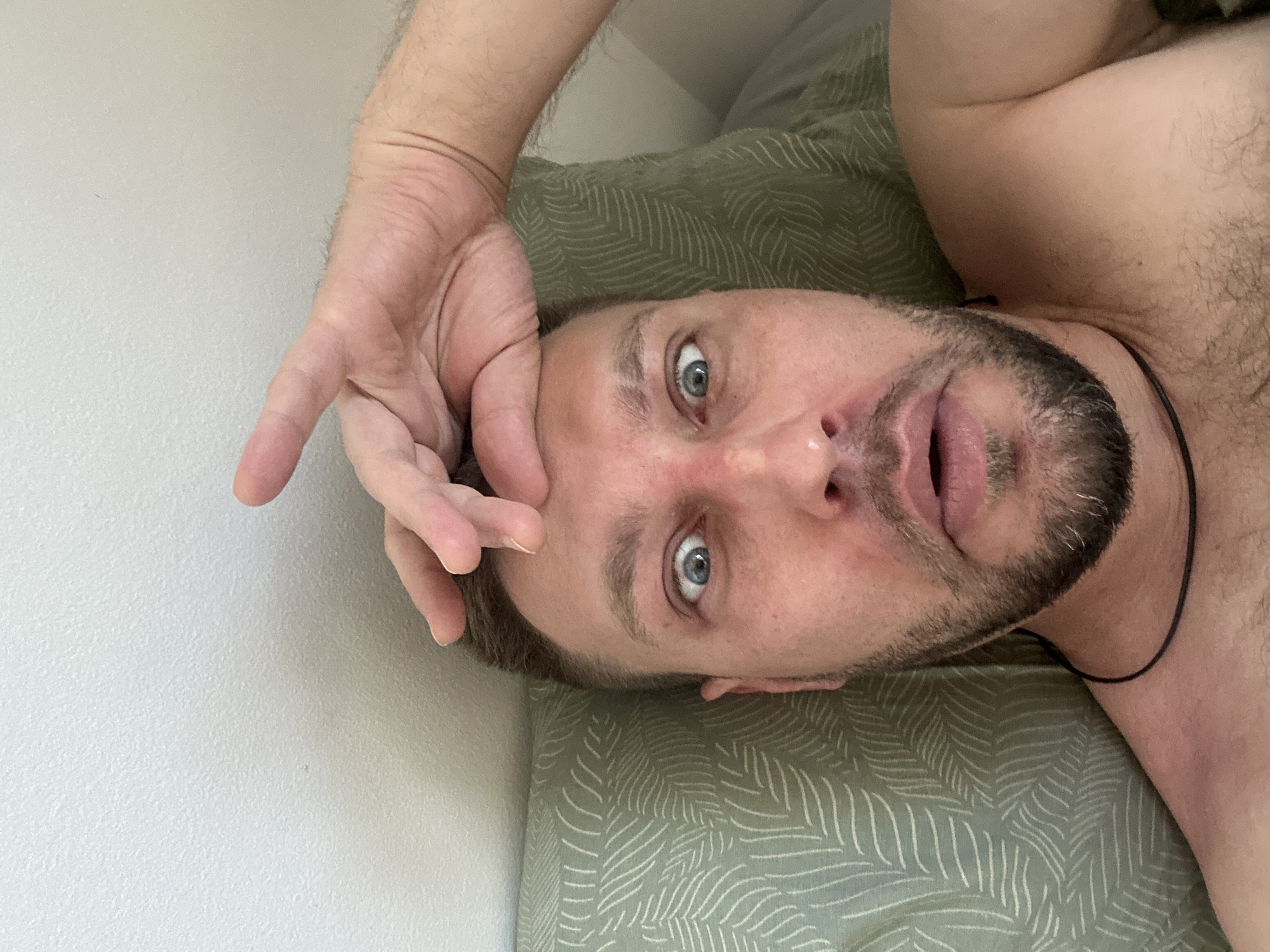 In a close-up, overhead shot, a fair-skinned man with blue eyes and a beard is lying down, looking directly at the camera with a surprised expression. His left hand is raised and touching his forehead. The man is wearing a black cord necklace. He is lying on a green pillow with a white leaf pattern. The background is a plain white wall. The scene appears to be indoors, likely in a bedroom, given the pillow and the man's relaxed posture. The lighting is natural, suggesting it may be daytime. No specific urban features or landmarks are visible to pinpoint a location within Zvezdara Urban Municipality, Serbia.