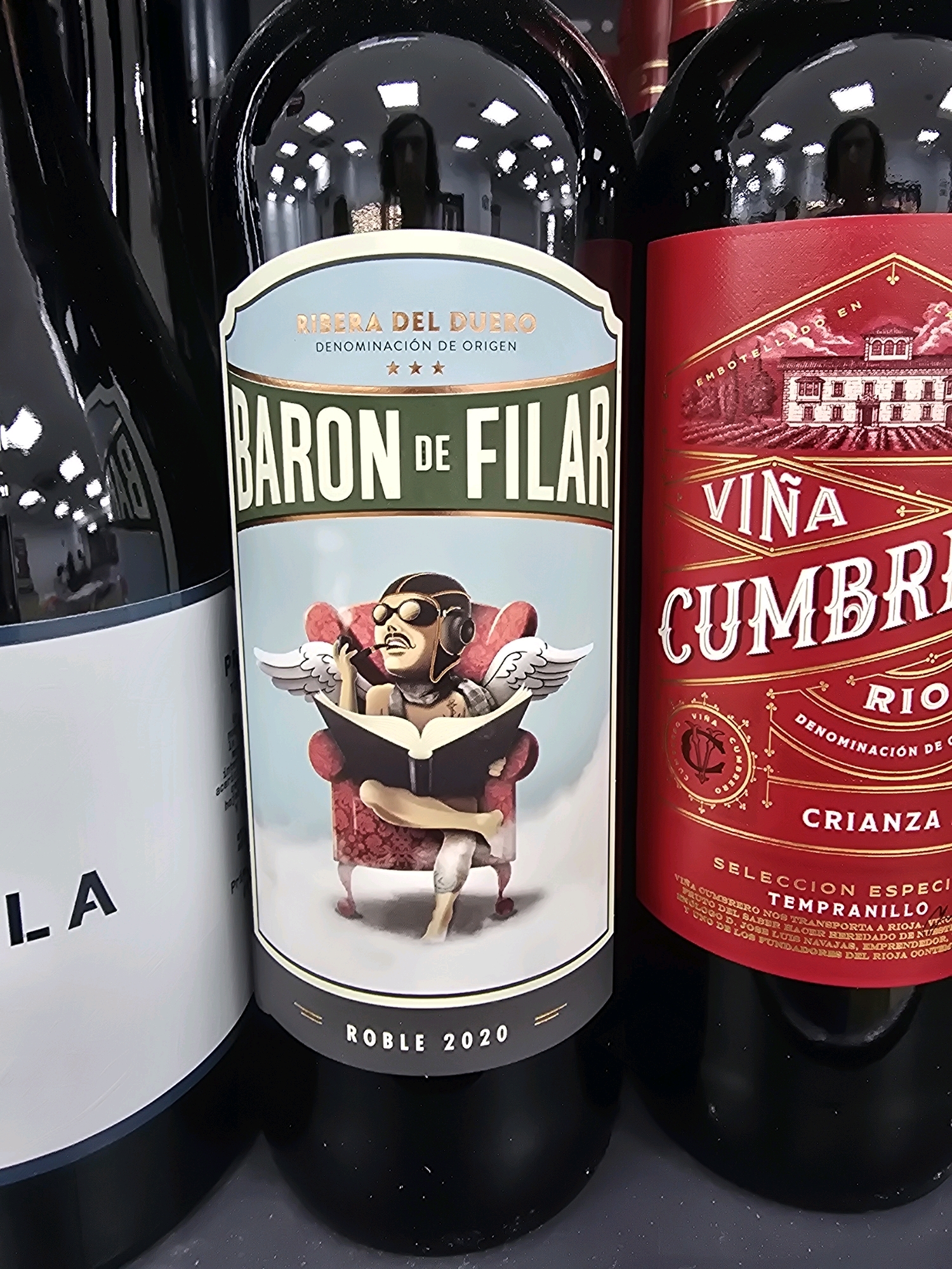 This is a close-up, eye-level shot of three bottles of red wine displayed side-by-side. The lighting is bright, reflecting off the glossy surfaces of the bottles, suggesting an indoor retail environment, possibly a wine shop or a supermarket aisle in Jaén, Spain.
The bottle on the left is partially visible, its label mostly obscured, showing only the word "LA" in black text against a white background.
The central bottle features a prominent label with a light blue background. At the top, it reads "RIBERA DEL DUERO" in a light brown, arched font, followed by "DENOMINACIÓN DE ORIGEN" and three small star symbols. Below this, the name "BARON DE FILAR" is displayed in large, green, block letters. The lower portion of this label depicts an illustration of a cartoonish character with goggles, a mustache, and wings, sitting in a red armchair and reading a book. The bottom of the label has "ROBLE 2020" in white text.
The bottle on the right has a vibrant red label with gold lettering. The top section features a drawing of a grand building with a red roof and white walls. Above this, in gold, is the text "EMBOTELLADO EN". Below the drawing, the label reads "VIÑA CUMBRERO" in large, ornate, gold lettering. Further down, it says "RIOJA" in gold, followed by "DENOMINACIÓN DE ORIGEN" and "CRIANZA" in gold. At the very bottom, in smaller gold text, are the words "SELECCION ESPECI TEMPRANILLO" and further descriptive text in Spanish that is mostly illegible due to resolution.
The bottles are arranged closely together, suggesting a curated display. The focus is sharpest on the labels of the central and right bottles.