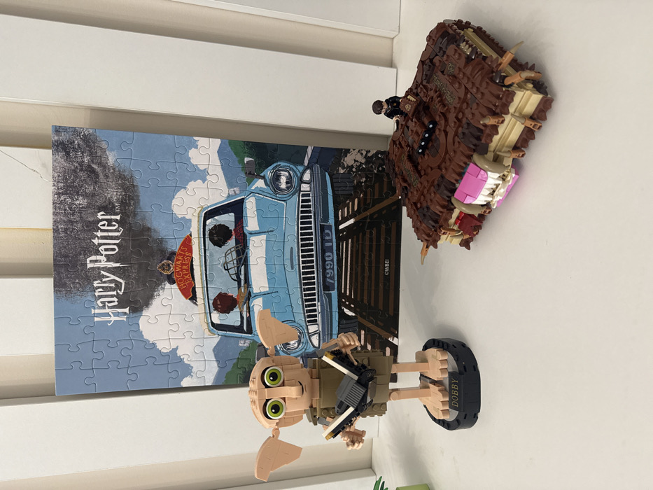 The scene, located in Batumi, Georgia, displays a collection of Harry Potter-themed items arranged on a flat, white surface against a white vertical paneling. Dominating the background is a completed jigsaw puzzle, depicting a light blue Ford Anglia flying low over railway tracks, with two figures (Harry Potter and Ron Weasley) visible inside. Above the car, a train labeled "Hogwarts Express" emits dark smoke. The puzzle is titled "Harry Potter" at the top and marked "©WBEI" at the bottom. To the left in the foreground stands a tan-colored LEGO buildable figure of Dobby the House-Elf, featuring prominent green eyes and holding a small, dark rectangular object in its hands. Dobby is positioned on a black circular base inscribed with "DOBBY". To the right, a brown LEGO model of the Monster Book of Monsters is present, characterized by its textured surface, exposed white and pink teeth, and multiple brown spikes. A small LEGO minifigure, dressed in dark clothing, is situated on top of the book.