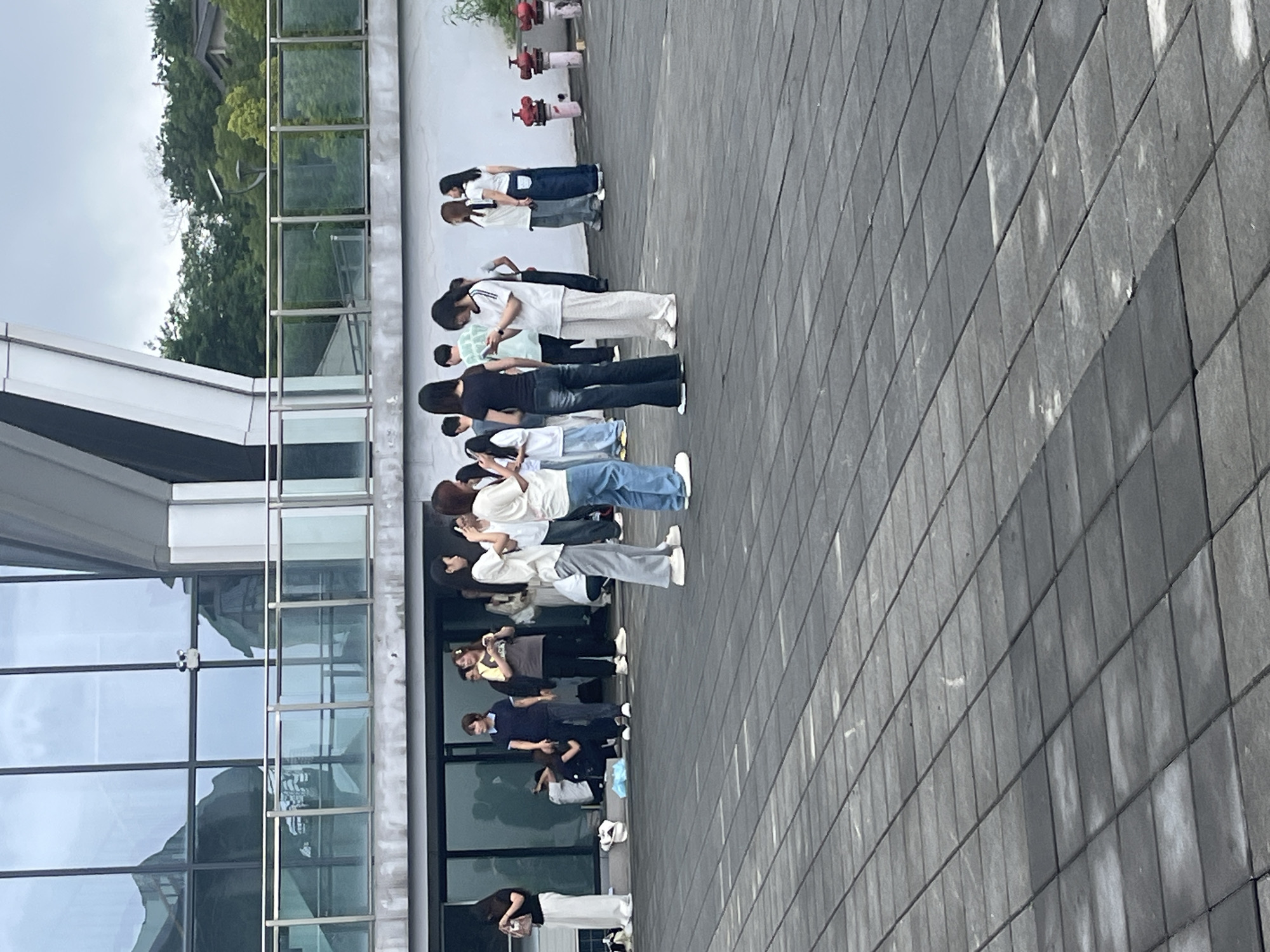 A group of young adults, appearing to be teenagers or in their early twenties, are standing on a paved outdoor area in front of a modern building. The building features large glass windows and a white facade with a metal railing. Some individuals are looking at their phones, while others are engaged in conversation. The paved area is composed of dark grey rectangular tiles. In the background, there are trees and a cloudy sky. Red fire hydrants are visible near the building on the right side of the image. The scene suggests a casual gathering or a pause between activities, possibly at an educational institution or a public space. The specific location in Jiangjin, China, is not evident from the visual details provided in the image.