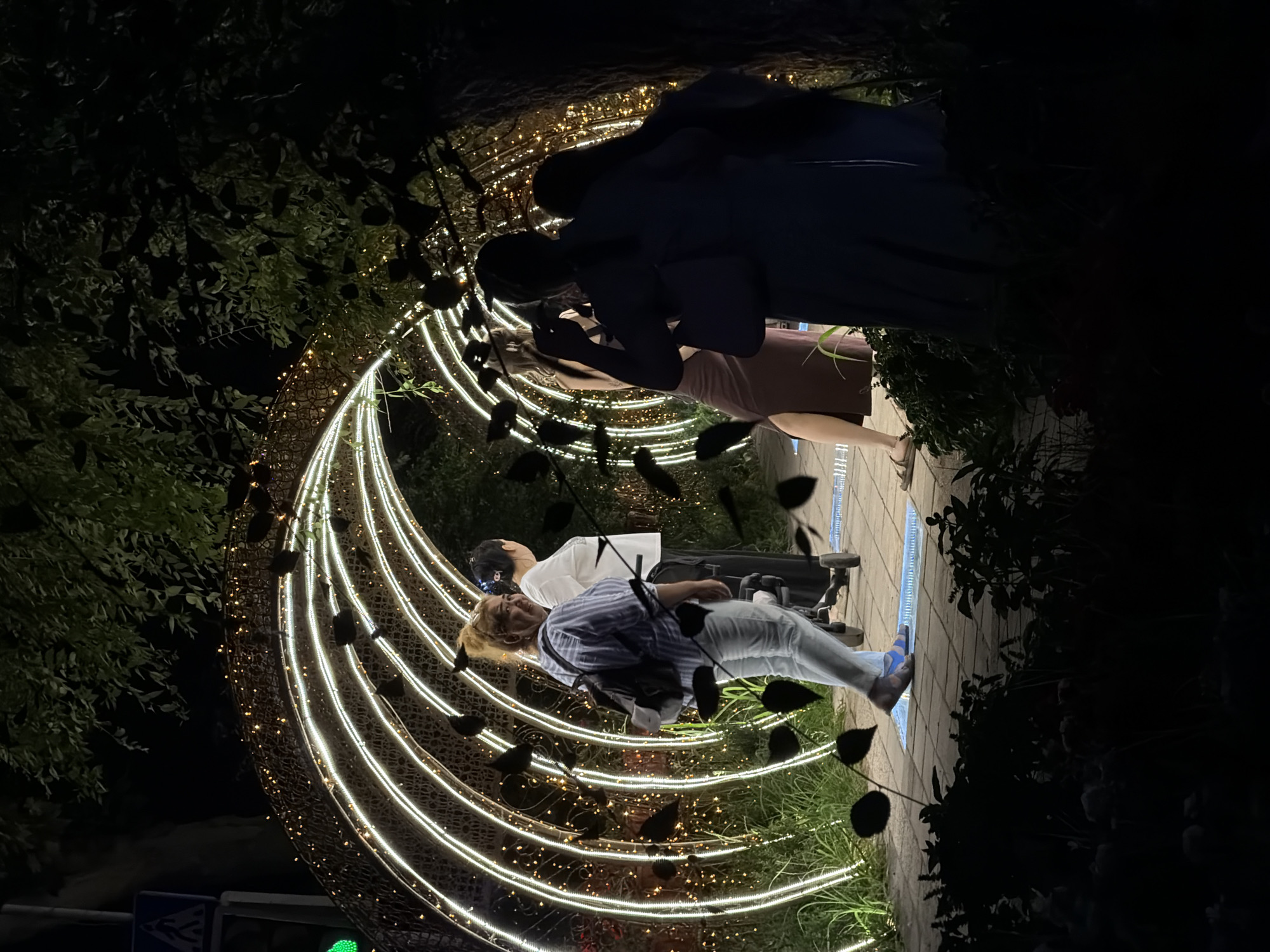 The image depicts a nighttime scene in a city park, possibly in Chirchik, Uzbekistan, featuring an illuminated archway decorated with white string lights and smaller yellow lights. Several individuals are present, some walking through or near the arch. A woman in the foreground, facing away from the camera, wears a blue and white striped shirt, light-colored pants, and blue sandals, carrying a backpack. To her left, another woman in a white shirt and dark pants pushes a stroller. Further into the arch, two figures are silhouetted against the lights, one appearing to hold up a phone, possibly taking a photo or video. The ground is paved with interlocking bricks, and lush green foliage surrounds the illuminated structure, casting shadows in the low light. Black leaf-shaped cutouts are suspended in the air around the archway, adding a decorative element. A street sign with a pedestrian icon is visible in the upper left corner, hinting at the proximity of a road or intersection.