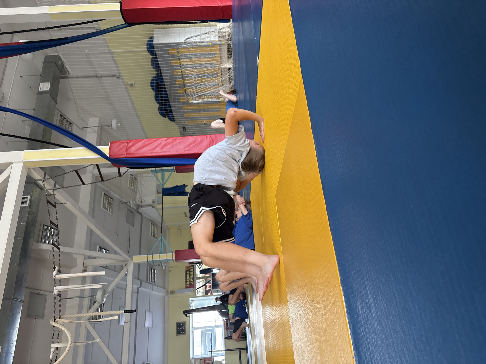 The image depicts an indoor gymnasium where several young boys are engaged in physical training on a yellow and blue mat. The central focus is a boy performing a backbend, with his body arched upwards, supported by his hands and feet. Other boys are lying on their backs or in similar resting positions in the background. The gymnasium appears to be equipped for gymnastics or martial arts, with climbing ropes, padded structures, and a sports net visible. The environment suggests an organized training session. The location is identified as the city of Chaikovsky, Russia.