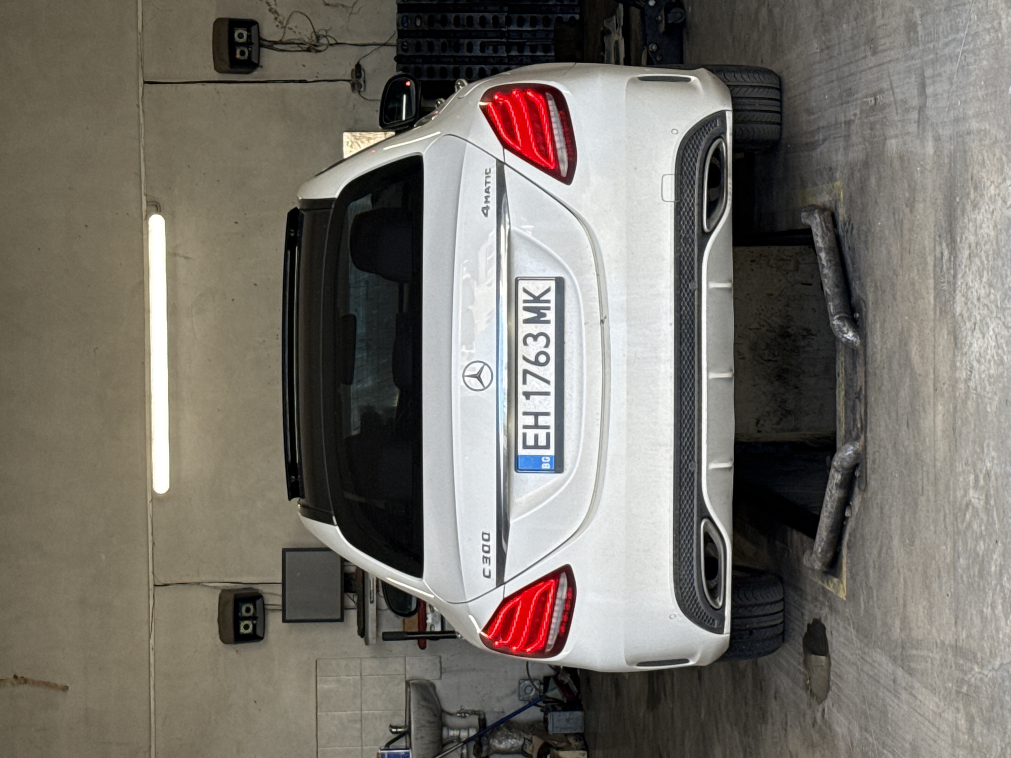 A white Mercedes-Benz C300 4MATIC is parked on a vehicle lift in a garage. The license plate reads "EH1763MK" with a "BG" symbol indicating registration in Bulgaria. The location is in Pleven, Bulgaria. The garage has concrete walls and floor. There is a fluorescent light on the left wall and electrical boxes mounted on the wall behind the car. Tools and equipment are scattered around the floor.