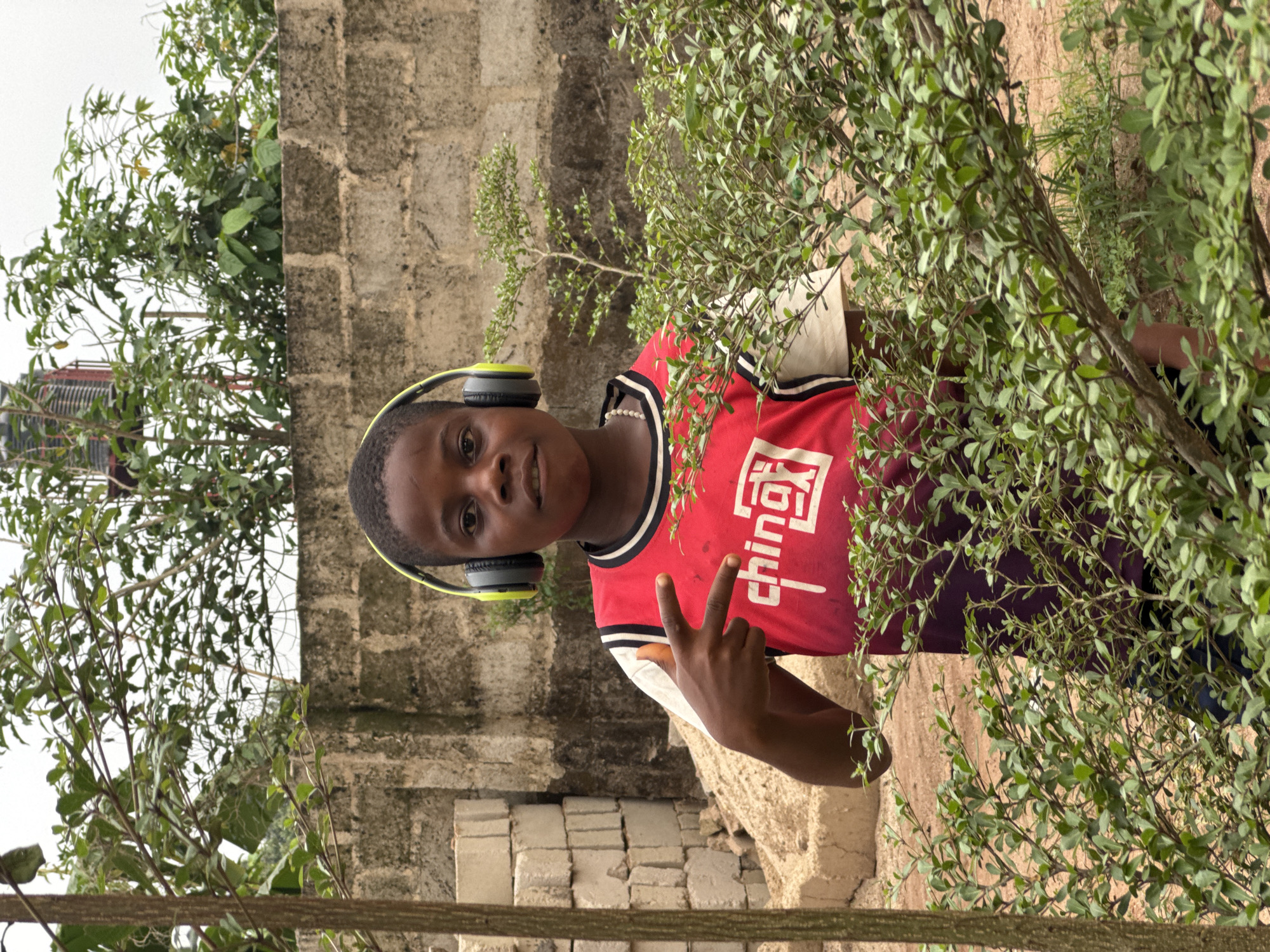 A young person is captured outdoors in Eziudo, Nigeria, positioned slightly right of the frame's center. The individual, appearing to be a boy, smiles cheerfully at the camera while making a peace sign with his left hand. He wears a red and white sleeveless jersey adorned with white Chinese characters '中国' (Zhōngguó), meaning 'China.' Lime green and black headphones are worn over his ears.
The scene is set amidst natural vegetation. Dense green bushes and leafy branches partially frame the subject, extending into the foreground on the right and background on the left. Behind the person, an unplastered concrete block wall provides a rugged backdrop. The ground appears earthy, possibly sandy. A bright, overcast sky is visible in the upper left, suggesting it is daytime. The atmosphere of the photograph is casual and portrays a moment of youthful ease.