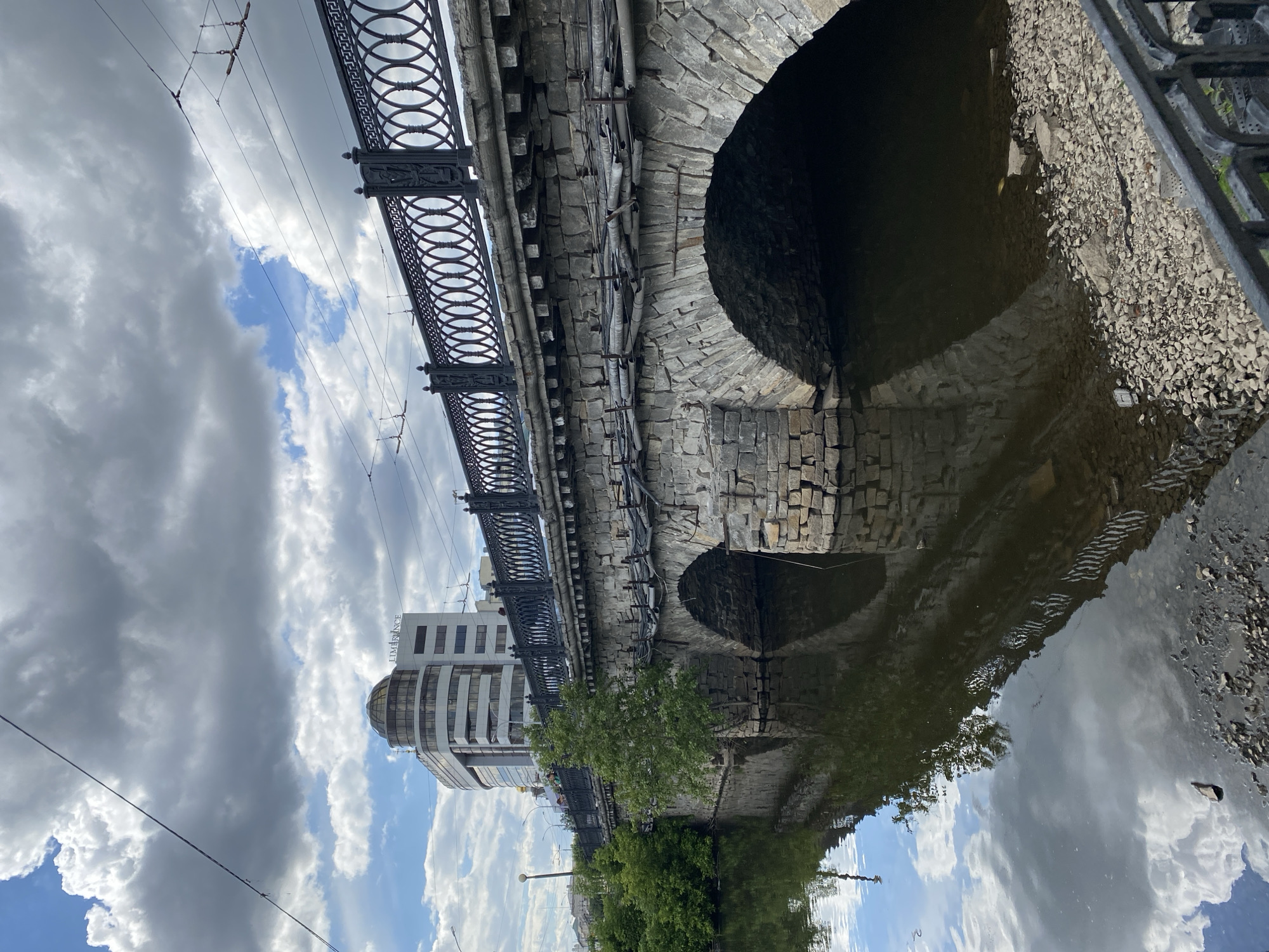 The image depicts a bridge over water in Yekaterinburg, Russia, featuring an arched stone structure reflected in the water's surface. The bridge has decorative metal railings. Overhead tram or electrical lines are visible against a cloudy sky. A building with a domed roof is situated near the bridge. A tree grows along the water's edge, its reflection also visible. The water's edge is made of stones and gravel.