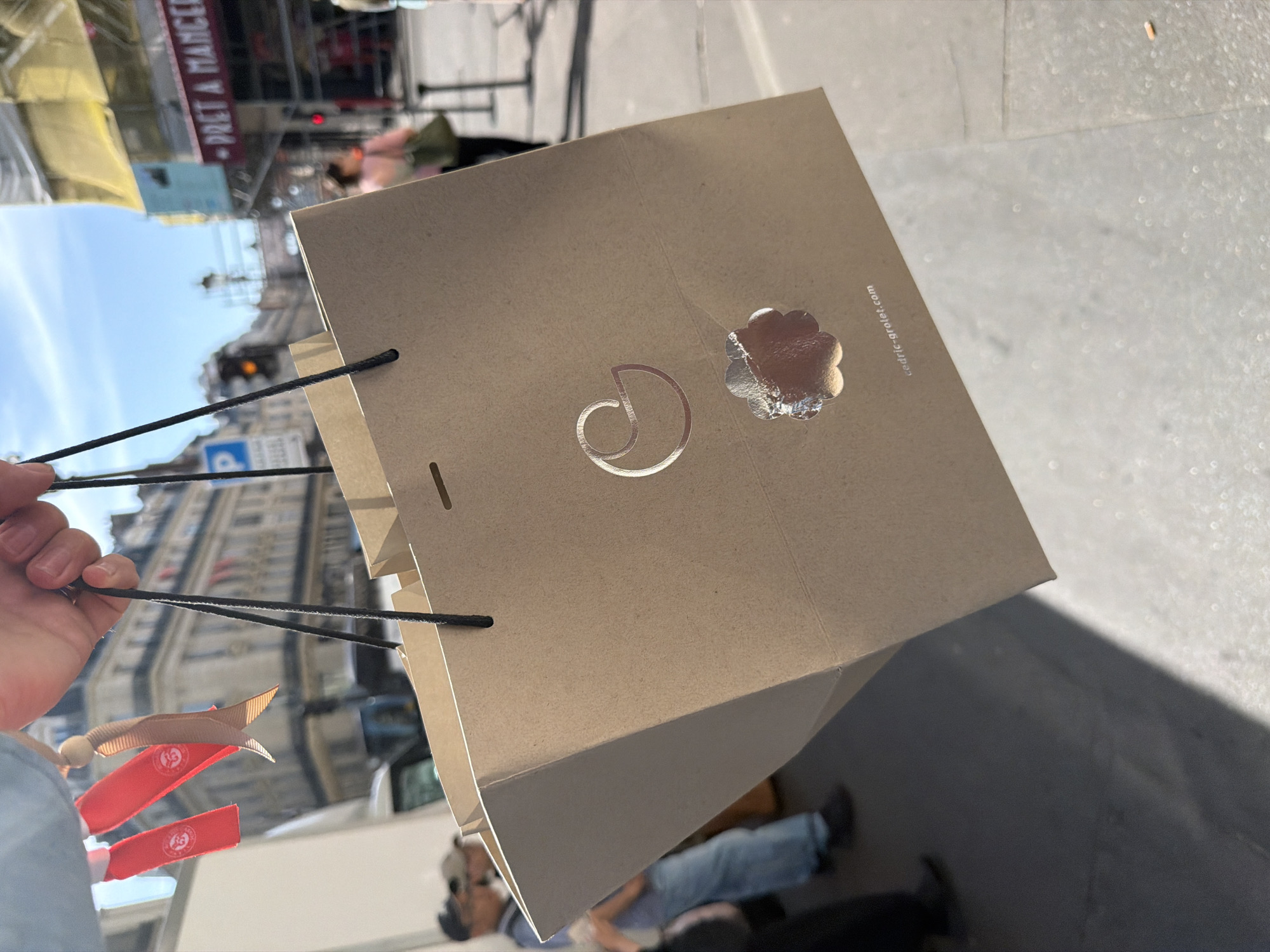 A person holds a tan shopping bag bearing a silver-colored logo of a stylized letter "D" next to a silver-colored flower-like design. The website address "cedric-grolet.com" is printed on the bag. The bag's handles are black cords. Attached to one of the handles are several Roland Garros-branded ribbons. The scene appears to be outdoors on a sunny day in Paris, France, as the background features Parisian architecture, including a building with a "PRET A MANGER" sign. Several people can be seen in the blurred background, and a shadow is cast on the ground.