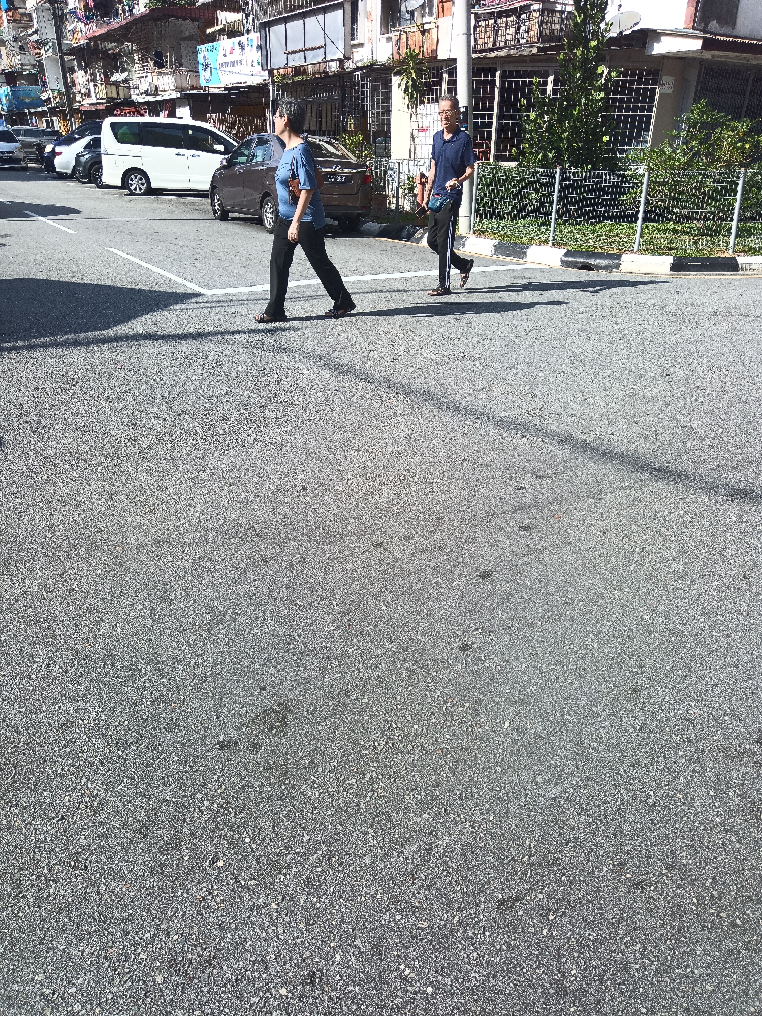 Two individuals are walking across a pedestrian crossing on a city street in Kuala Lumpur, Malaysia. The person in the foreground is a woman wearing a blue t-shirt and black pants, walking from left to right. The person behind her is a man in a blue polo shirt and black pants with white stripes, also walking from left to right. Several vehicles, including a white van and a dark-colored car, are visible in the background, parked or driving on the street. The buildings lining the street appear to be multi-story structures, some with signs and balconies. The ground is paved with asphalt, marked with white lines indicating the pedestrian crossing. The scene is captured during daylight, with shadows cast on the road, suggesting a sunny day. The environment is urban, characterized by buildings, vehicles, and street infrastructure.