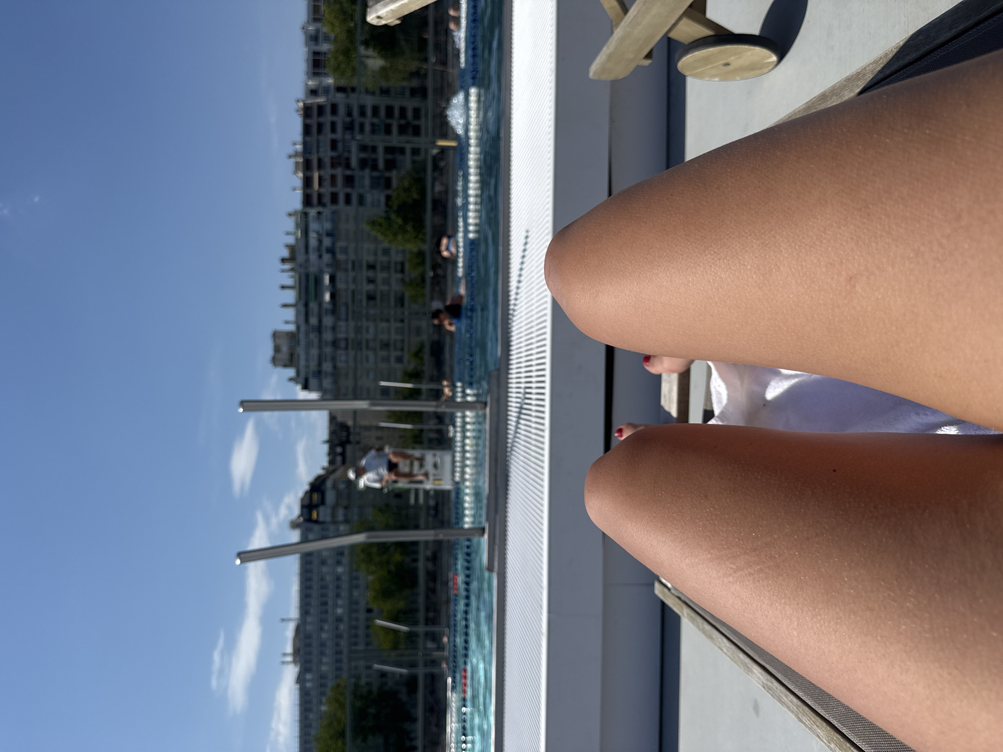 The image shows a first-person perspective of someone relaxing on a lounge chair. In the foreground, the legs of the person are visible, tanned and smooth with goosebumps. A white towel is placed on the lounge chair. In the mid-ground, there is a swimming pool with people swimming and a lifeguard on a high chair. The background shows a row of tall buildings, characteristic of the Parisian cityscape. This scene is likely at a public pool or a rooftop pool in Paris, France, possibly on a sunny day given the bright sky and clear weather.