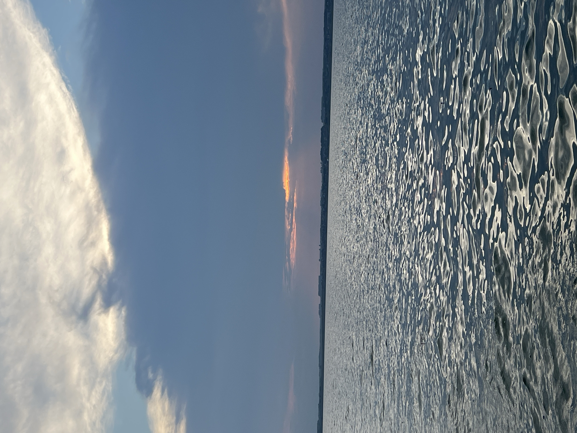 The image depicts a serene body of water, likely a lake or reservoir, under a dramatic sky. The water surface is rippled, reflecting the muted light from the clouds and the horizon. In the distance, a faint line of land is visible, dotted with what appear to be buildings, suggesting a settlement or town along the shoreline. The sky is a mix of deep blues and grays, with patches of lighter clouds illuminated by sunlight breaking through, creating streaks of orange and yellow. The overall impression is one of twilight or a stormy sunset. The location is specified as Novopichugovo, Russia.