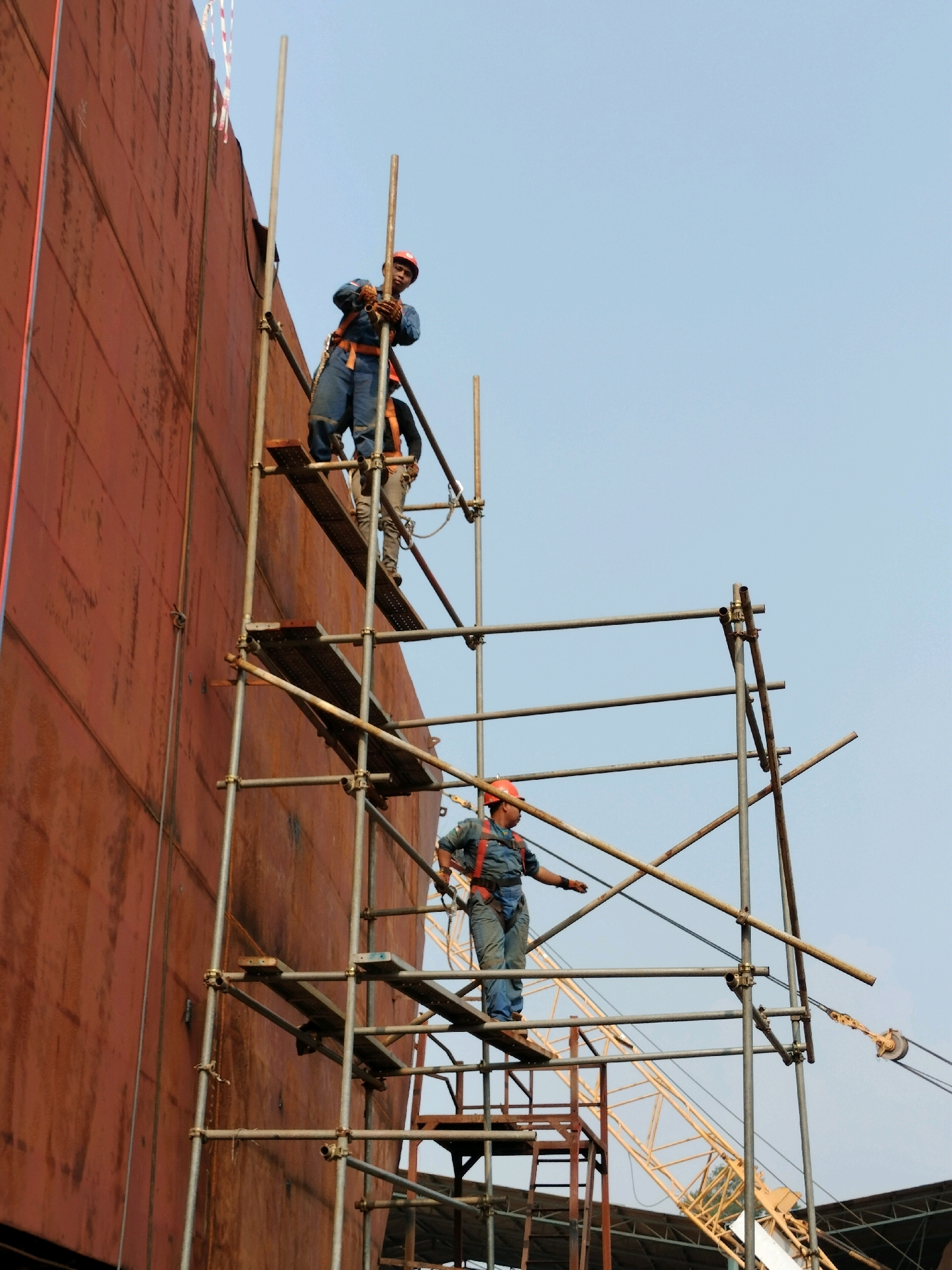 In Batam, Indonesia, three construction workers are visible on scaffolding attached to a large, rust-colored vessel, likely a ship or industrial structure. Two workers are higher up on the scaffolding, one at the top actively holding a metal pole, and another slightly below. A third worker is positioned on a lower platform, looking to the left. All three are wearing blue overalls and orange safety helmets, and at least two are secured with safety harnesses. The scaffolding is made of metal poles and planks. In the background, a yellow crane and parts of a covered structure are visible against a clear blue sky.