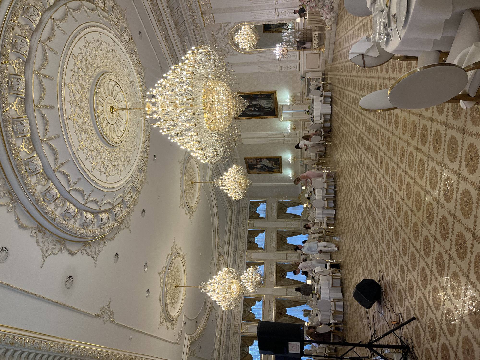 The image depicts an opulent indoor event hall, likely in Dubivtsi, Ukraine, set up for a formal gathering or banquet. The scene is dominated by elaborate gold and white decor, featuring ornate ceiling moldings, large circular medallions, and grand crystal chandeliers that illuminate the space with a warm glow. The tables are draped in white linen and set with formal place settings, suggesting a meal or reception is in progress.

Several individuals are visible, dressed in light-colored attire, seemingly attending to guests or working at the event. They are positioned around the tables and along the periphery of the hall. The atmosphere appears elegant and celebratory. Large framed paintings adorn the walls, adding to the refined ambiance. The floor is covered in a patterned tile. In the foreground, sound equipment, including a speaker and cables, is visible, indicating that music or speeches might be part of the event. The lighting suggests it is either evening or an indoor setting that is artificially lit. The overall impression is one of a luxurious and well-attended occasion.