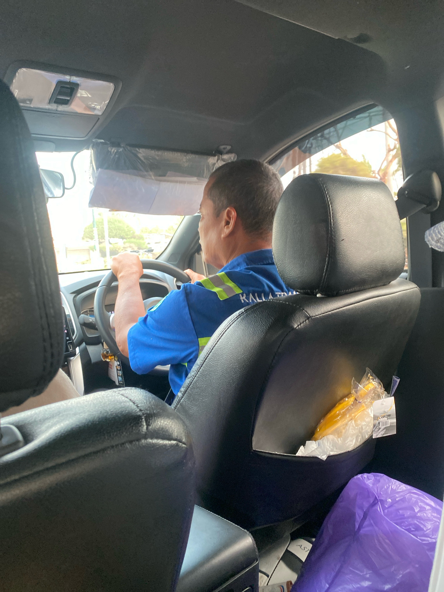 An exterior shot of a black vehicle's interior in Banjarbaru, Indonesia, shows a man driving. The man, wearing a blue uniform with reflective stripes, is viewed from behind the passenger seat, his hands on the steering wheel. The vehicle's interior includes black leather seats, a sun visor with plastic covering, and a dashboard. A purple plastic bag and a yellow object wrapped in plastic are visible on the passenger seat. Outside the vehicle, trees and buildings are visible under a bright sky.