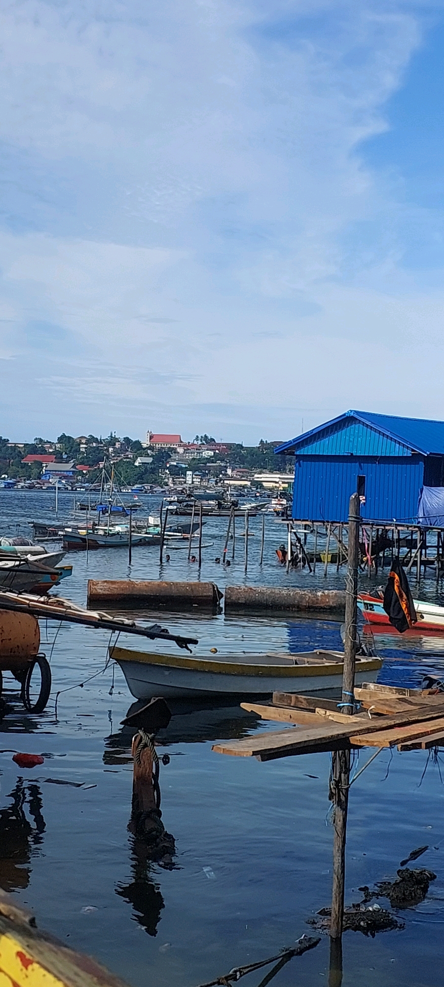 This is a vertical, medium shot of a waterfront scene in Jayapura, Indonesia, likely in the area of Skyland or Holtekamp, given the characteristic stilted housing and the bay. The image depicts a calm, blue body of water filled with various boats and floating structures. In the foreground, a light-colored boat with a yellow trim rests on the water, tethered to a wooden post. To the left, a wheelbarrow with orange parts is partially visible. Several large, cylindrical objects, possibly buoys or barrels, are submerged in the water.
The middle ground shows a collection of smaller boats, some appearing to be fishing vessels, and a larger, bright blue building constructed on stilts with a blue tarp extension. Several people are visible in the water near the blue building, some appearing to swim. A dark garment with an orange emblem hangs from a wooden pole near the blue building.
In the background, the city of Jayapura stretches across a hilly landscape. Buildings of various sizes and colors are clustered together, with a prominent white building with a red roof, possibly a church or government building, situated on a higher elevation. The sky is a pale blue with scattered white clouds. The overall impression is one of a bustling, working waterfront in a tropical coastal city.