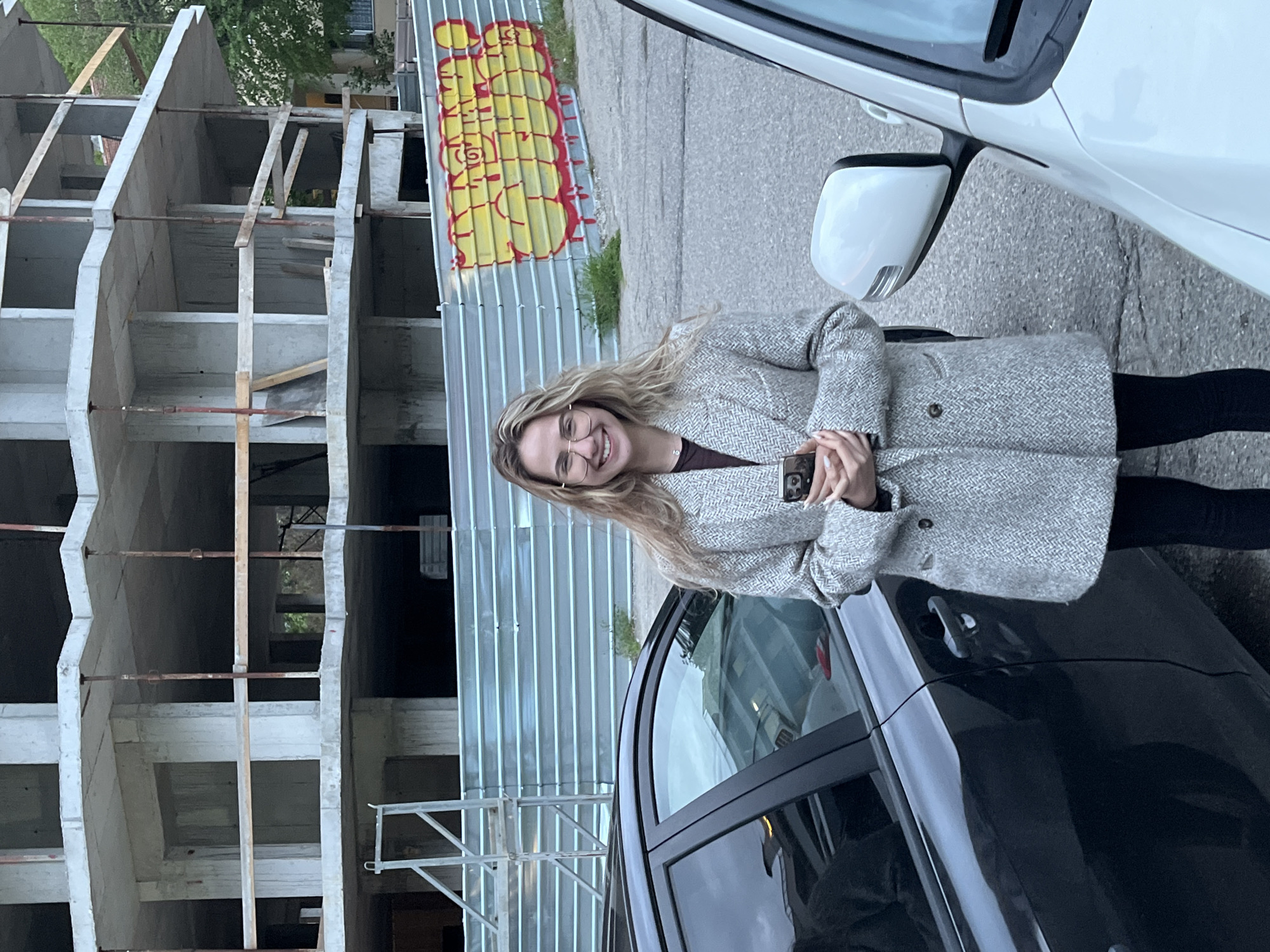 A woman is standing between two cars in front of a construction site and a corrugated metal fence. The woman has long blonde hair, wears glasses, a grey herringbone coat, black leggings, and is holding a smartphone. To her left is a partially constructed concrete building with scaffolding. Behind her is a corrugated metal fence with graffiti on it. To her right are two cars, one white and one black. The location is Blagoevgrad, Bulgaria.