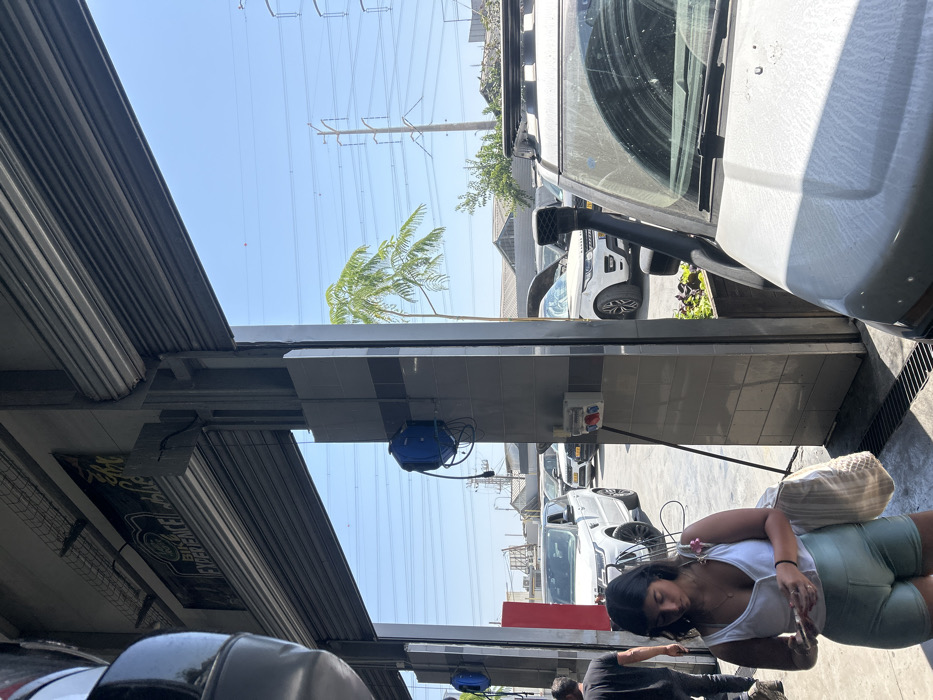 A woman in a white tank top and green shorts stands next to a car wash station, holding a phone. Several vehicles are parked in the background, including a white Land Rover. Overhead, utility poles stretch into the distance against a clear sky. Part of a building's awning and support structure frames the scene. A blue hose reel is mounted on the tiled wall of the car wash, near a power outlet.