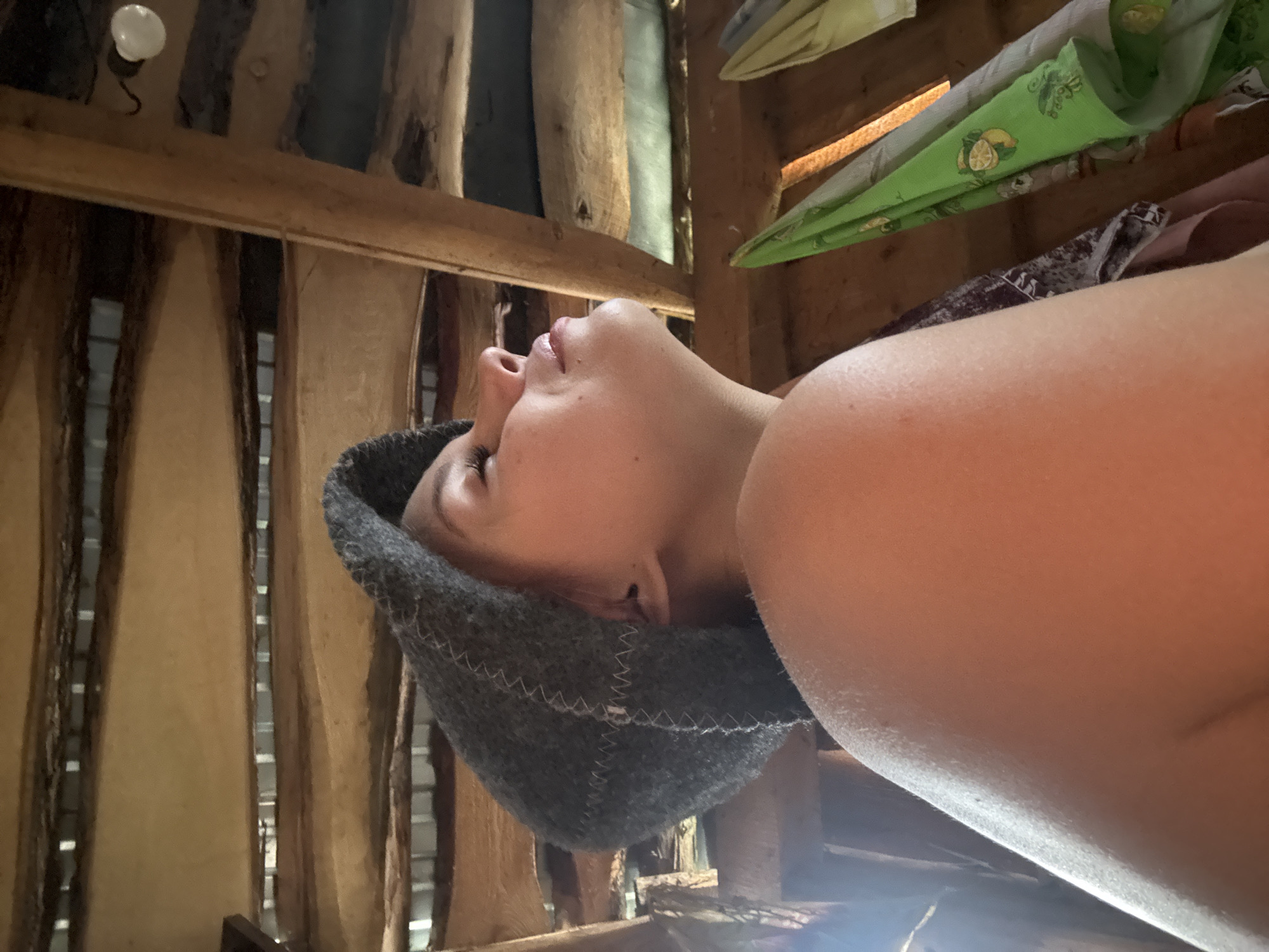 The image shows a close-up, angled view of a person relaxing in what appears to be a rustic wooden interior, likely a sauna or banya, in Chaikovsky, Russia. The person, whose profile (nose, lips, chin, and part of an eye) is visible, is wearing a grey felt hat, commonly used in saunas to protect from heat. Their eyes are closed, and their expression suggests calm and relaxation.
The background consists of rough-hewn wooden planks and beams, forming the walls and ceiling of the structure. Some gaps between the planks allow glimpses of a darker area beyond. A simple, white light bulb hangs from a wire in the upper left corner. In the upper right, several folded textiles are placed on a wooden shelf or beam; one is distinctly green with a yellow lemon pattern, while others are muted colors. A warm light highlights the person's arm and shoulder, indicating a light source from below or the side, possibly reflecting off the skin. The overall setting is simple, practical, and functional, consistent with a traditional bathhouse environment. No discernible text is visible in the image.