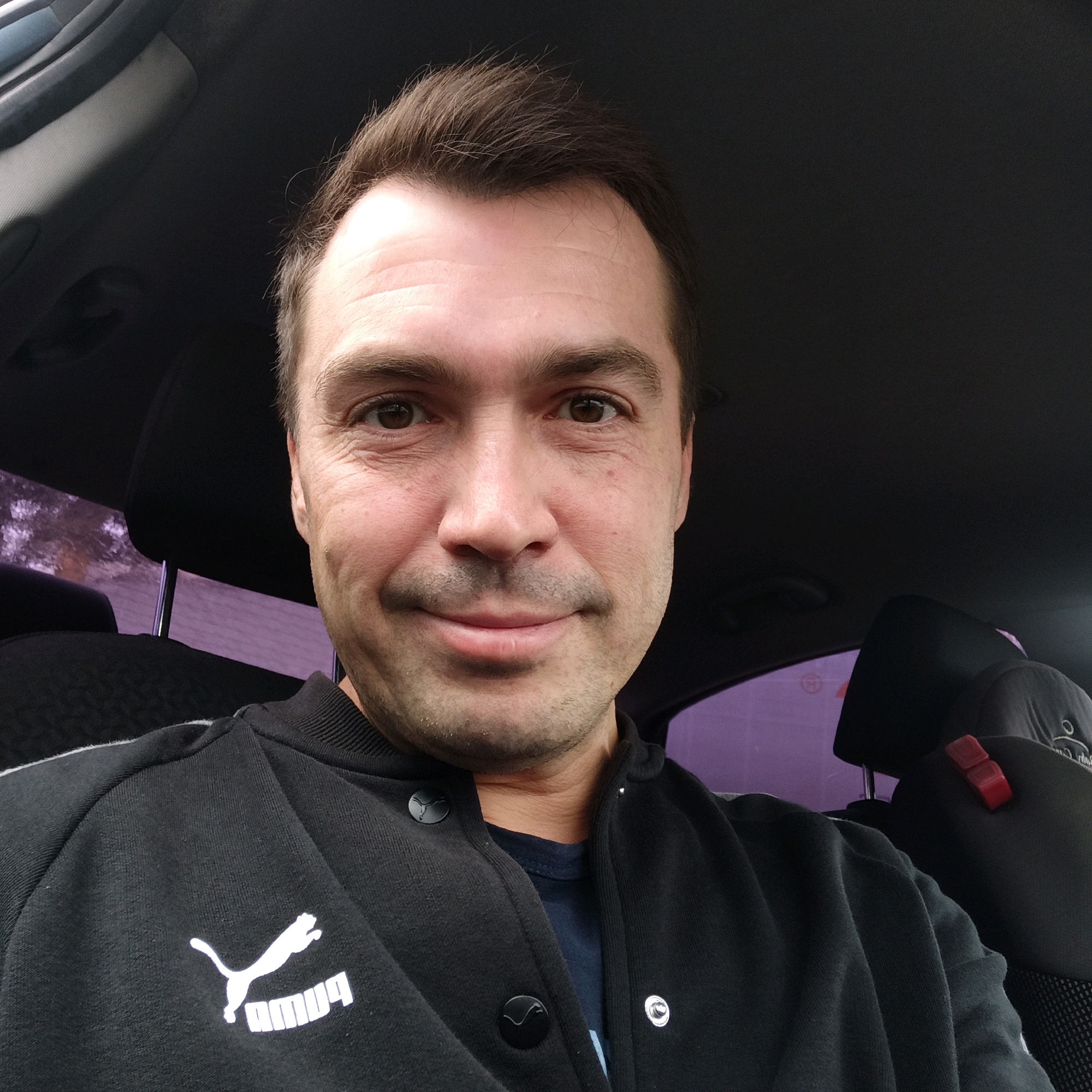 A medium shot photograph captures a man in his 30s, with brown hair styled upwards and subtle stubble, sitting in the driver's seat of a car. He is wearing a black Puma jacket with the brand's logo visible on the left chest. The man is smiling faintly and looking directly at the camera. The interior of the car is dimly lit, with the right side of the frame showing a headrest and a portion of the car's window, tinted purple, through which faint foliage is visible. The background is dark, suggesting the photo was taken indoors or at dusk. The overall impression is that of a casual selfie taken during a car ride. The specific location within Nizhny Novgorod, Russia, cannot be determined from the provided image.