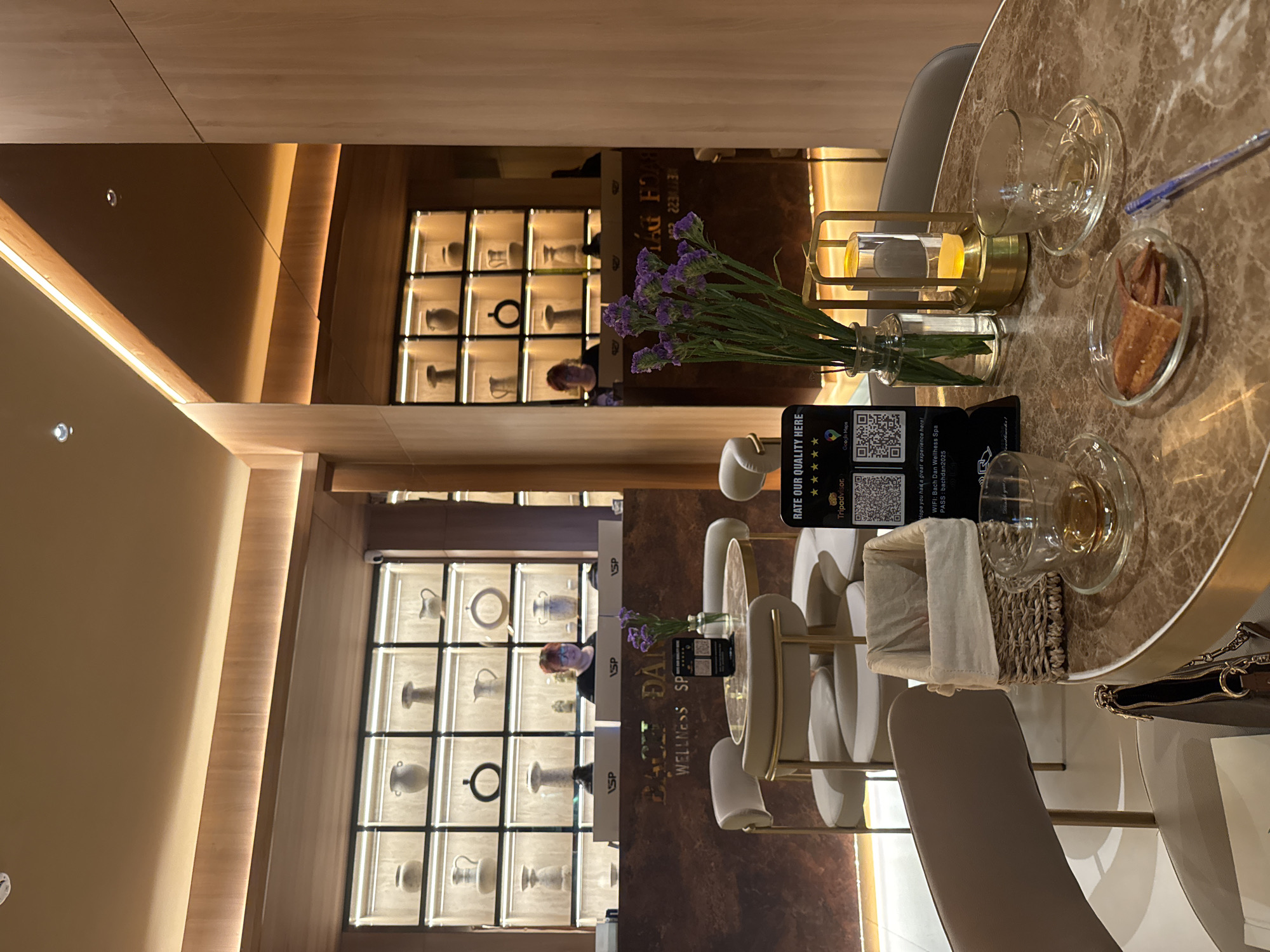 Here is a detailed description of the image:
The image depicts a cozy and aesthetically pleasing interior space, possibly in Unknown, Unknown. Dominating the scene is a round, marble-patterned table set for two, with clear cups and saucers, a small plate of food, and a decorative lantern-like holder with a candle. A small vase with purple flowers adds a touch of color to the arrangement.
A prominent black sign is placed on the table, featuring a QR code and the text "RATE OUR QUALITY HERE," suggesting a business is soliciting customer feedback. The sign also includes a five-star rating system, Tripadvisor, and Google Maps logos.
The chairs are modern, upholstered in a light color that matches the overall aesthetic. There's a glimpse of a handbag, indicating someone has recently been seated or is about to be.
In the background, a series of backlit display cases are visible, each containing what appear to be decorative pottery or ceramic items. The lighting is warm and inviting, with the use of wood tones on the walls and trim.
A mirror effect is present, which creates a sense of depth and doubles the elements of the room, including the display cases. A person with red hair is vaguely reflected in the mirrored surface.
The environment suggests a wellness center, spa, or a high-end lounge area, indicated by the sign that reads "BACH DAN WELLNESS SPA," which is also displayed on the lower portion of the background.