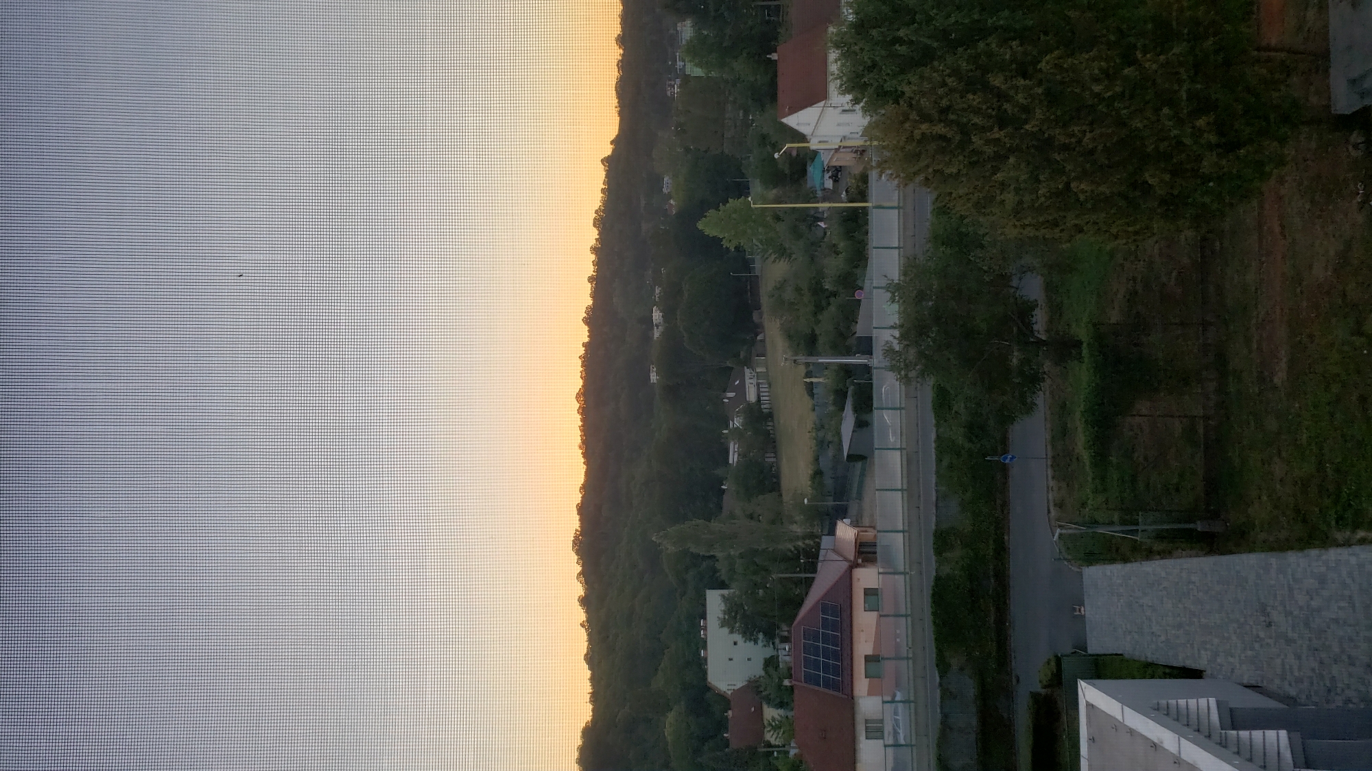 The image captures a view of a suburban area during a sunrise or sunset. The foreground shows the edge of a modern building with a light-colored exterior and a paved walkway leading away from it. A chain-link fence runs along the side of the walkway and extends into the middle ground, separating it from a grassy area with trees.
The middle ground features a road with a few cars visible and a blue circular sign with a white arrow on it. Beyond the road, there is a cluster of houses, one with solar panels on its roof. Further in the background, a tree-lined hill rises, with scattered houses and structures visible among the trees. The sky above transitions from a pale blue at the top to a warm orange glow near the horizon, indicating a sunrise or sunset.
Based on the distinct architecture of the houses, particularly the one with solar panels and the overall suburban layout, and considering the potential for such a view within Prešov, Slovakia, this scene is likely located in a residential area on the outskirts of the city, possibly in a neighborhood like Sídlisko III or a similar development characterized by a mix of single-family homes and apartment buildings, with nearby green spaces and hills. The specific location is likely an elevated viewpoint, perhaps from an apartment building or a higher floor of a residential complex, looking out towards the east or west to capture the atmospheric lighting.
