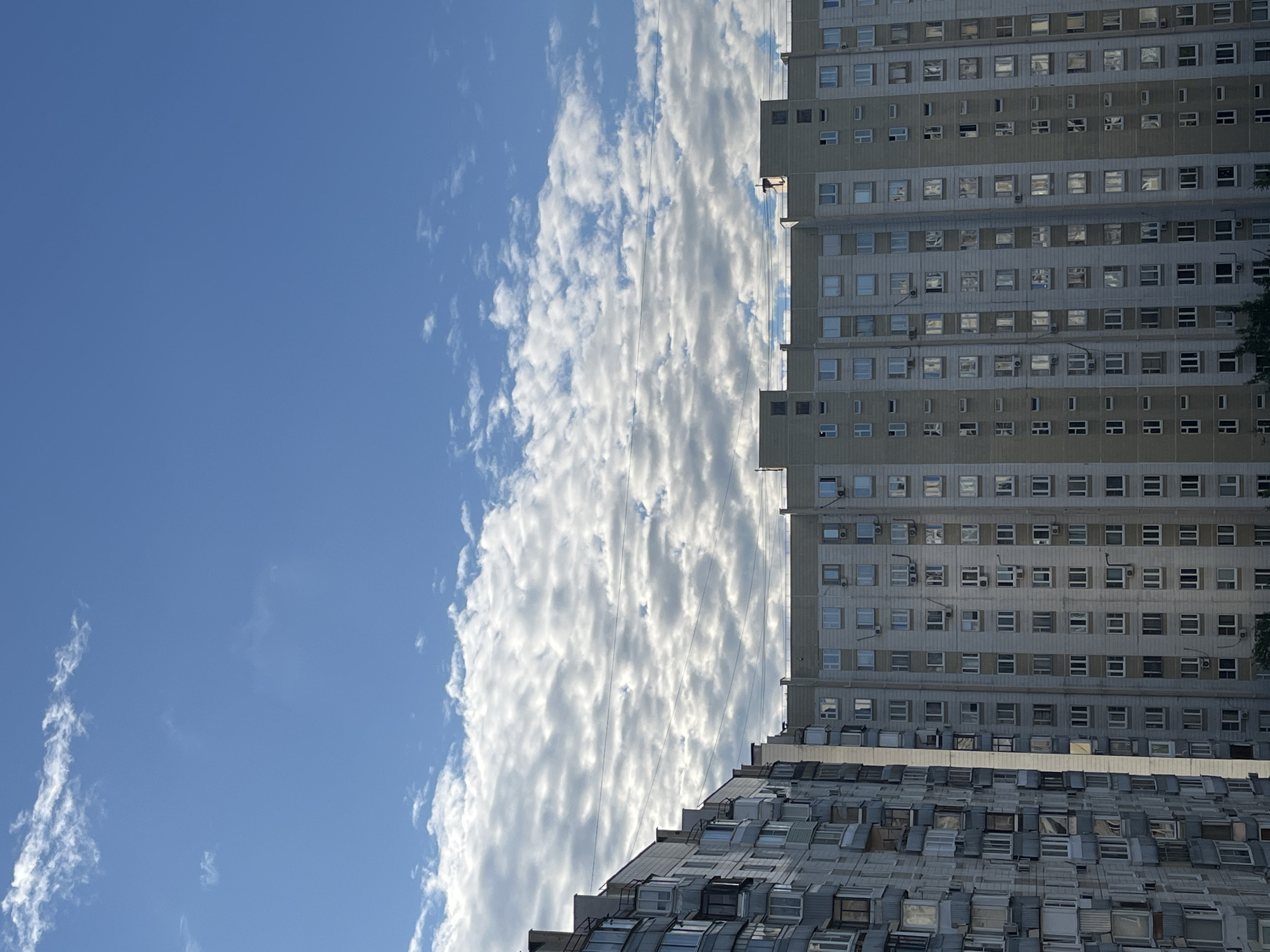 The image shows the facades of two large apartment buildings under a bright blue sky with scattered white clouds. The building on the left has a more varied facade with many balconies, some enclosed with windows. The building on the right is more uniform in its design, with rows of windows and air conditioning units. The scene appears to be in an urban residential area, likely a district of Moscow, Russia, given the architectural style of the buildings, which are typical of Soviet-era mass housing. There are no people visible in the image. The overall environment is daytime, with clear weather.