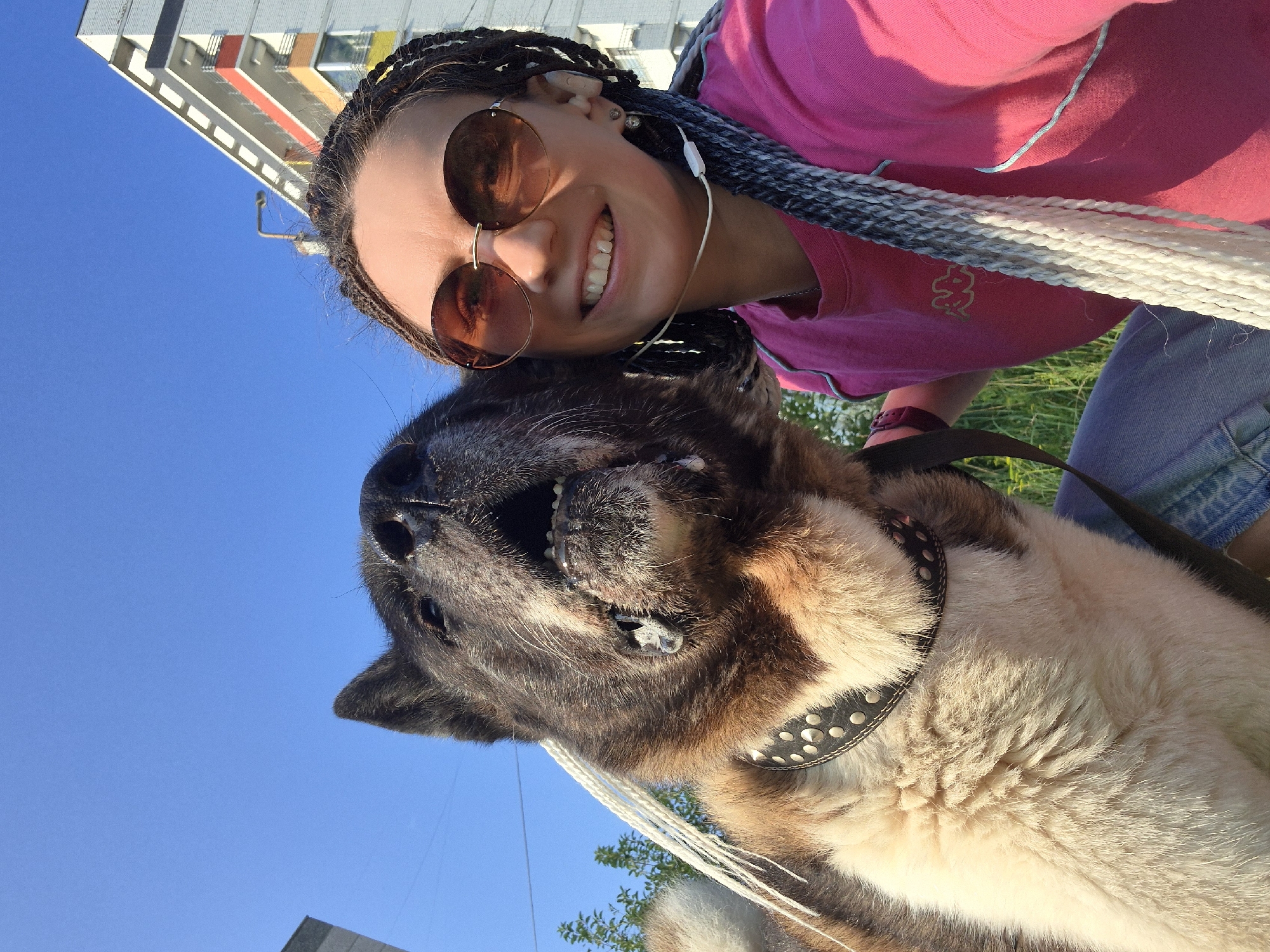 In a low-angle, selfie-style shot taken on a sunny day in Chaikovsky, Russia, a woman with long, braided hair and sunglasses smiles at the camera. She is wearing a pink t-shirt and jeans. Next to her, a large dog with dark fur and a studded collar is looking up, mouth open, with its tongue visible. In the background, a multi-story building with colorful balconies and a blue sky are visible. The setting appears to be an outdoor, urban environment.