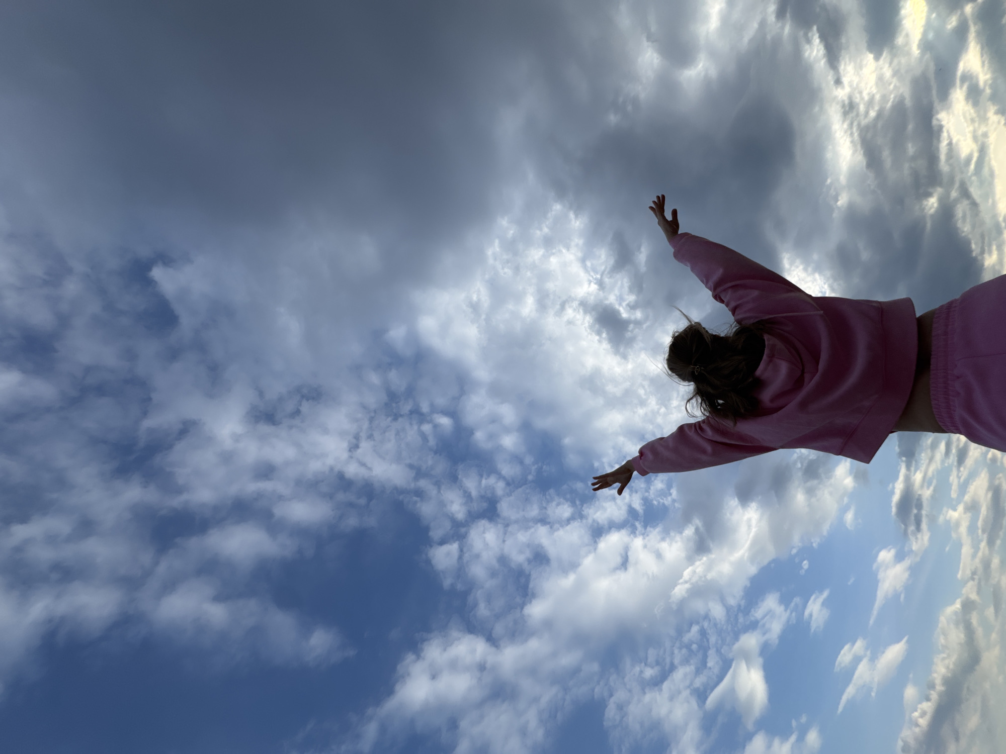 A low-angle shot shows a person in a pink sweatshirt and pants standing with arms outstretched against a cloudy sky. The sky transitions from dark grey clouds in the upper left to blue sky with white clouds in the lower right. The person's back is to the camera, with their head slightly tilted to the left. The location is Noviy, Russia.