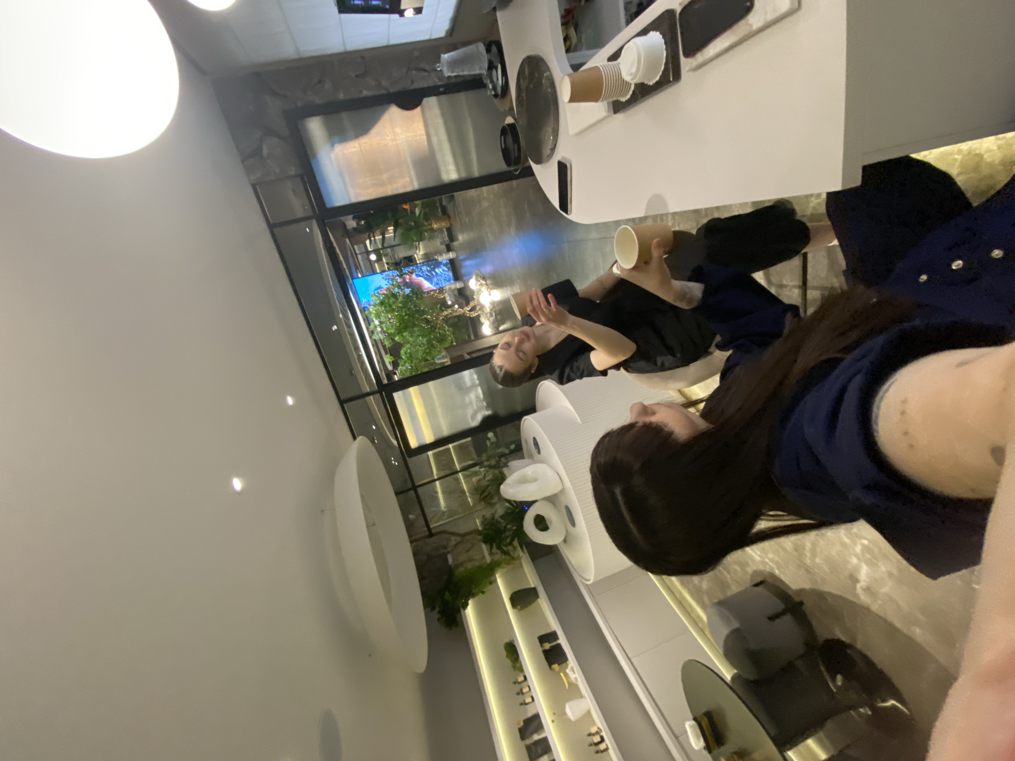 A horizontally oriented, high-angle photograph depicts the interior of a brightly lit establishment, likely a cafe or restaurant. Two individuals are visible: a man holding a cup, and a woman with dark hair wearing a blue garment. They are seated at a white table where multiple cups and a mobile phone are placed. An adjacent table is also white. A large window with a view of a street with vegetation is prominent. Various design elements, including a white circular fixture and shelving with displayed items, adorn the walls. The location is possibly in Votkinsk, Russia, but this cannot be forensically confirmed from the visual information provided.