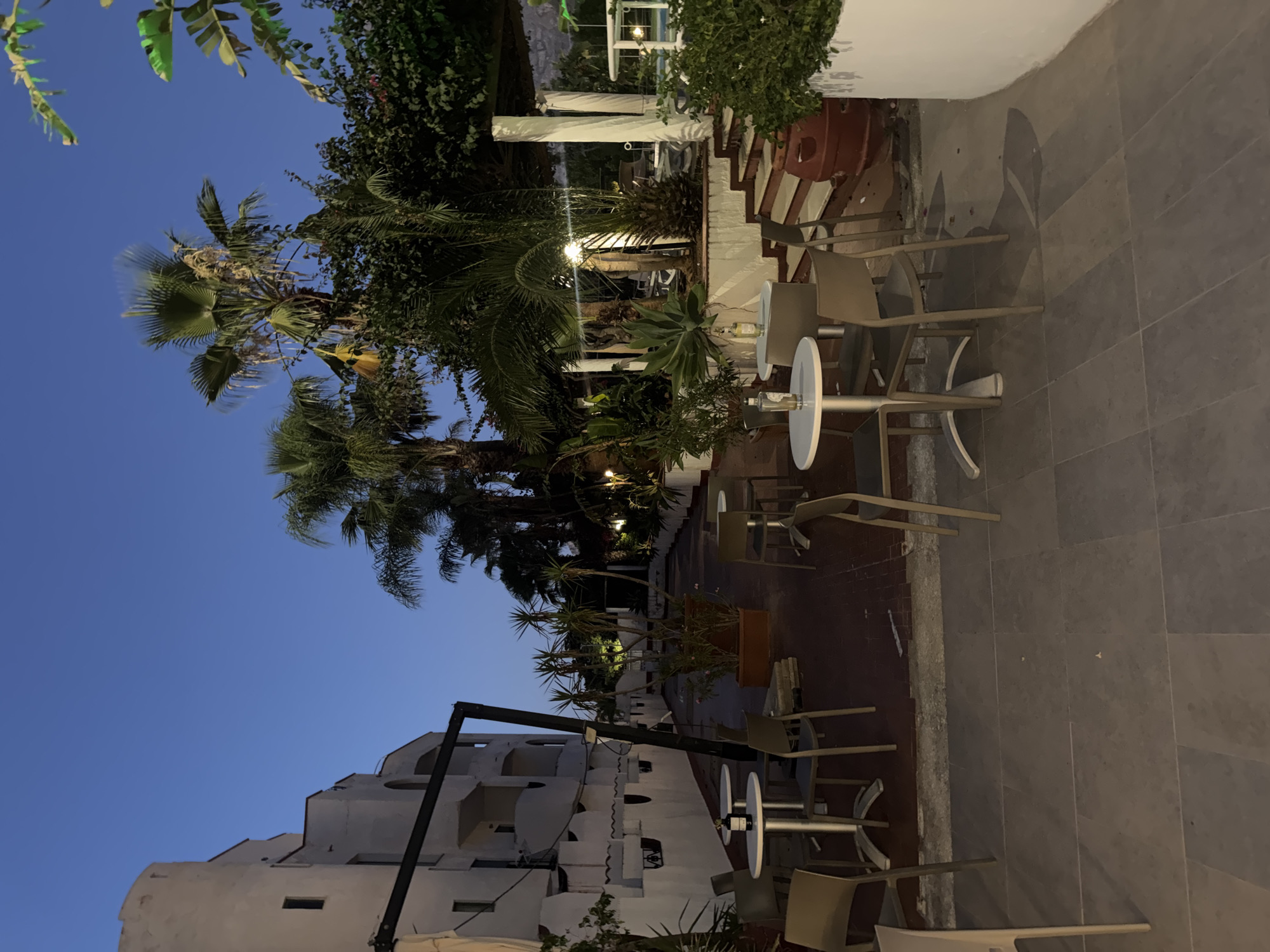 The image depicts an outdoor seating area at dusk, likely a restaurant or cafe. In the background, a white, multi-story building with arched windows is visible. Several palm trees and other lush greenery surround the area.
Scattered throughout the scene are several round tables with metal bases and chairs, some of which appear to be made of a light-colored material. On some tables, bottles of what appears to be wine or spirits and glasses are present, suggesting the area is set up for patrons.
The ground is paved with large, gray tiles in the foreground and a reddish-brown surface further back. A set of stairs with a white railing leads up to a covered area, also supported by white pillars.
The sky is a deep blue, indicating twilight. The lighting suggests artificial illumination from within the building and possibly from lamps in the seating area, casting shadows. The overall impression is of a serene evening setting.
The image is taken from a low-angle perspective, looking across the seating area towards the building and palm trees. There are no people visible in the frame. The location is identified as Isola delle Femmine, Italy.