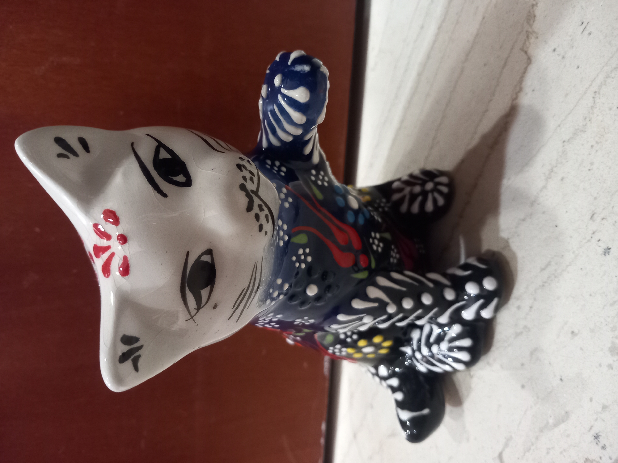 A ceramic cat figurine is seated on a light-colored surface with dark horizontal lines. The cat has a white head with black and red decorative markings. It is wearing a dark blue vest adorned with colorful floral patterns in red, yellow, green, and white. The cat's front right paw is raised. The background is a dark reddish-brown surface.