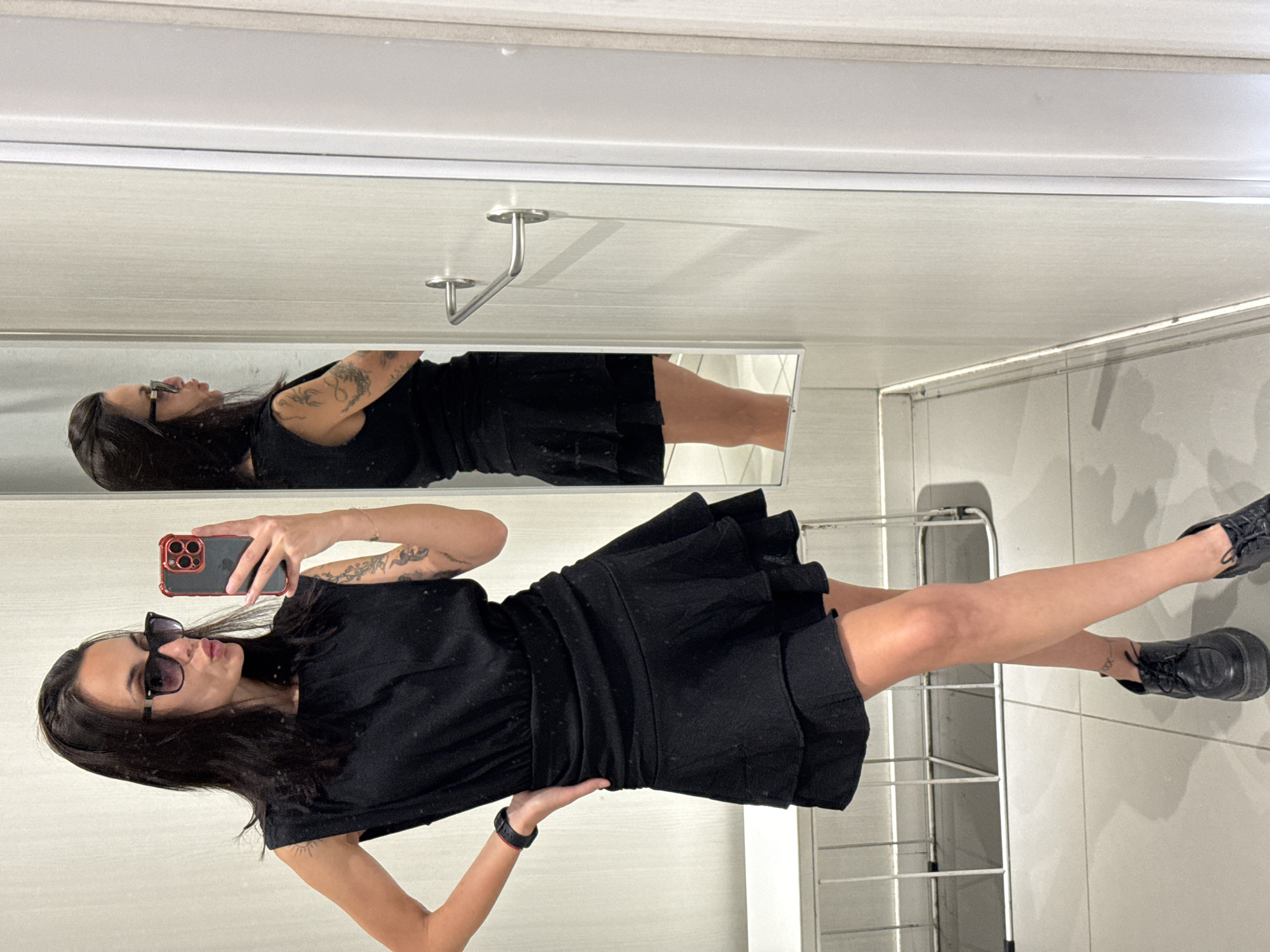 A woman is posing for a mirror selfie in what appears to be a dressing room or bathroom setting. The mirror captures her full-body reflection and a partial view of her face in the mirror. She is wearing a black dress and black shoes. Her hair is dark. She is holding a smartphone in her hand, presumably to take the picture. A towel rack is visible behind her. The location is indiscernible and cannot be conclusively connected to Pleven, Bulgaria, based on the provided image.