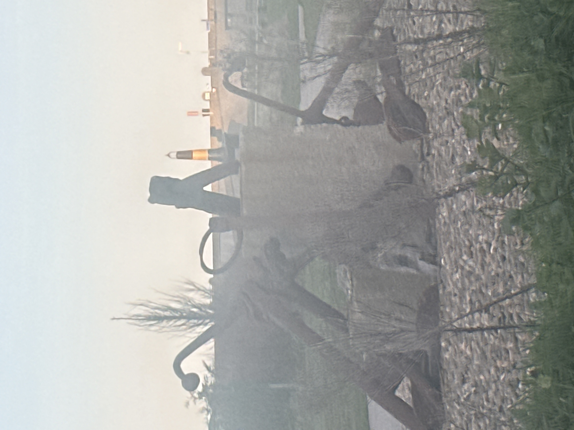 The image depicts a blurry outdoor scene in East Suffolk, United Kingdom. A metal structure, possibly an artistic installation or part of a playground, is prominently featured in the foreground. The ground appears to be covered in gravel or small stones. Vegetation is visible on the right side of the frame. In the background, indistinct buildings and streetlights are discernible, suggesting an urban or suburban setting. The image is heavily obscured by water droplets or mist, reducing clarity and detail.