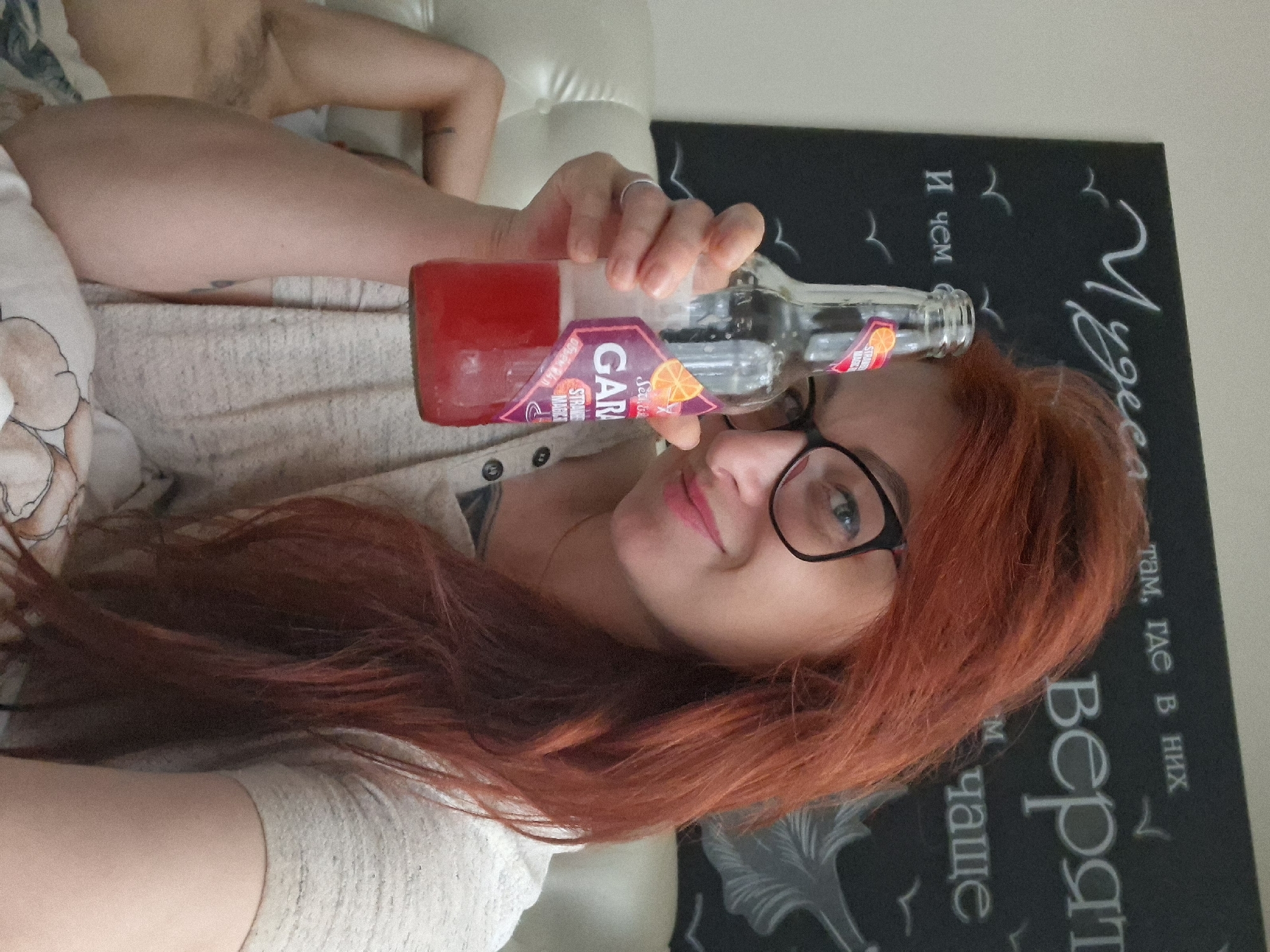 A woman with long, red hair and glasses holds a bottle of a red beverage in front of her face, looking at the camera. She is wearing a light-colored top. In the background, there is a dark chalkboard with white writing and drawings. The woman is reclining on a white surface that appears to be a bed. A portion of another person's arm and torso is visible on the left side of the image, also reclining. The image does not contain enough information to determine the location in the city of Myadzyel, Belarus.