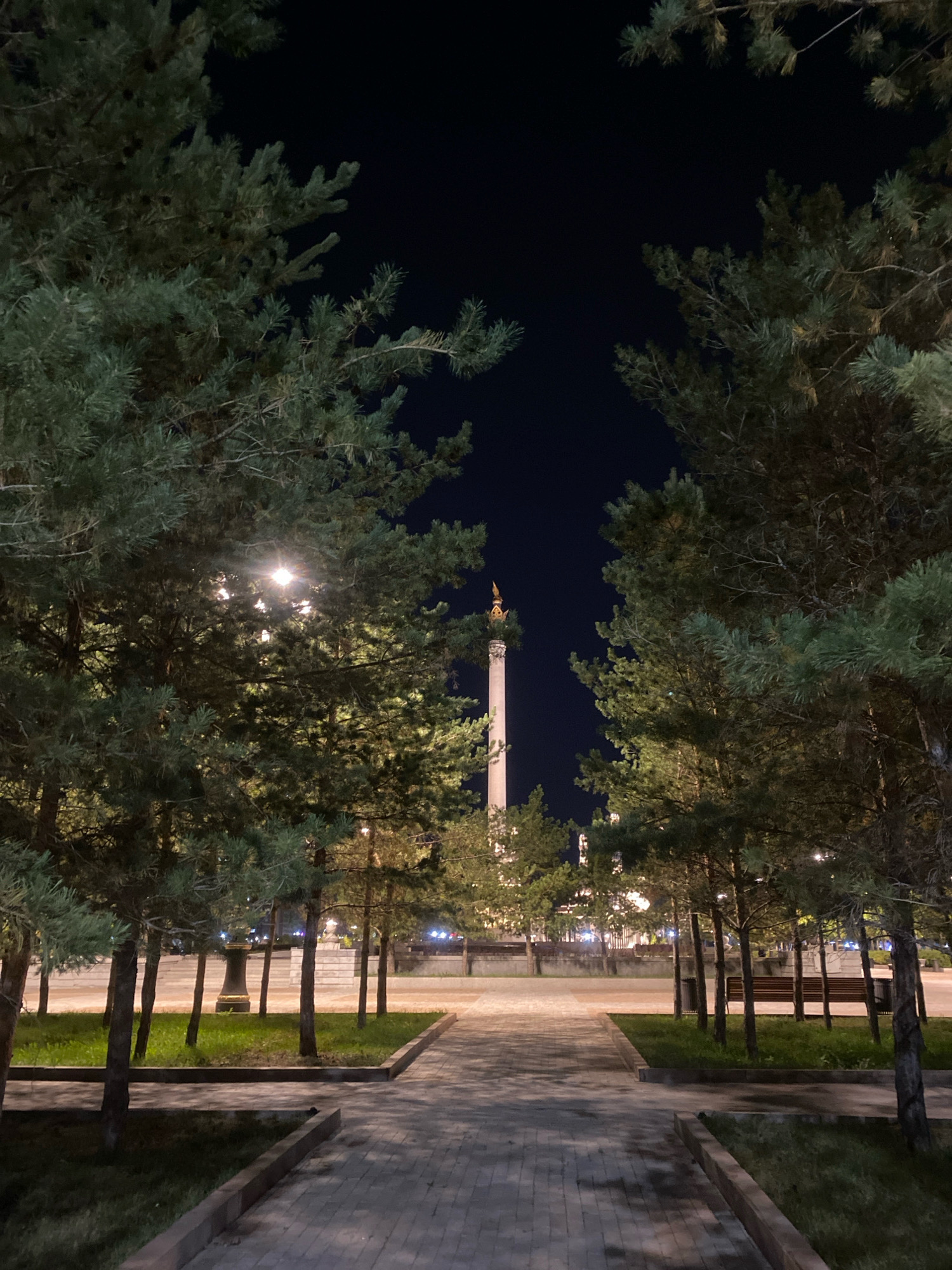 The image shows a pathway lined with trees leading towards a tall monument at night. The monument has a golden cap and is illuminated. Streetlights are visible through the trees. In the background, there are buildings with lights on. The pathway is paved with bricks and surrounded by grass and trees. The scene appears to be in a park or public space in Astana, Kazakhstan, likely the Monument of Independence.