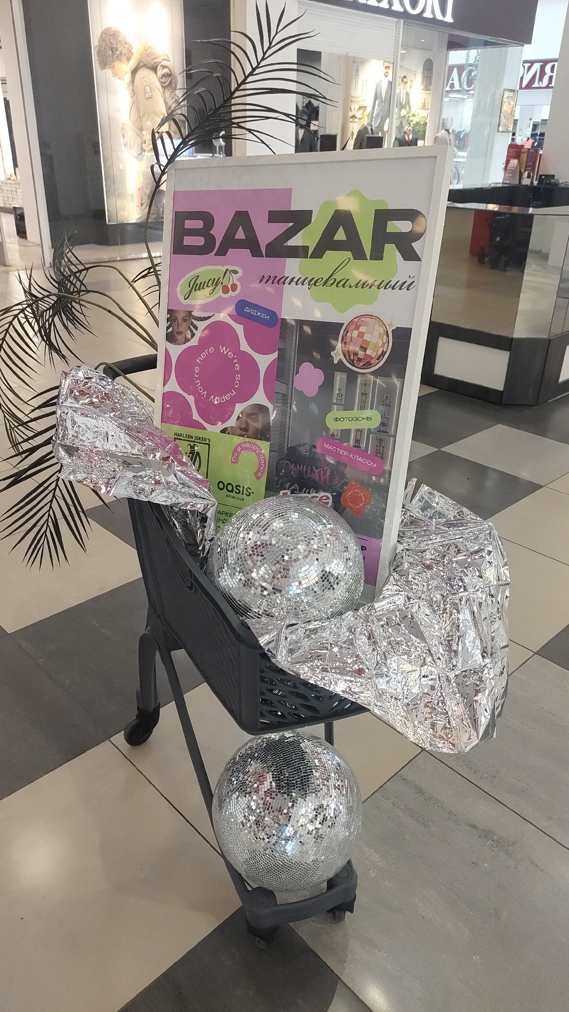 The image shows a display in a shopping mall, presumably in Unknown, Unknown. In the foreground, there is a shopping cart filled with crumpled silver foil, topped with a large disco ball. A second disco ball sits below the cart on the floor. Inside the cart, there is a white-framed poster with the word "BAZAR" written in large, bold letters, in pink and green. The word "танцевальный" is written below "BAZAR" in a smaller font. The poster is adorned with stickers of various designs, including images of celebrities and event details. Behind the shopping cart, there is a large potted plant with black palm-like leaves. In the background, there are several storefronts with mannequins dressed in various outfits. The floor is tiled with a geometric pattern. The overall scene has a promotional or decorative appearance.