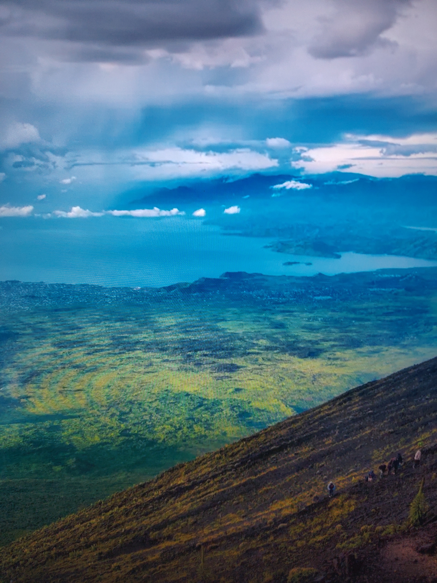 The image shows a landscape with a mountain slope in the foreground. Several people are scattered along the slope, appearing to be hiking or working on the hillside. The terrain is a mix of dark soil and vegetation. In the midground, there is a green expanse that transitions into a body of water under a cloudy sky. The sky is filled with various cloud formations, ranging from dark, heavy clouds to lighter, scattered ones. The overall scene suggests a view from a high elevation overlooking a landscape with diverse natural elements. The location of this image is unknown.