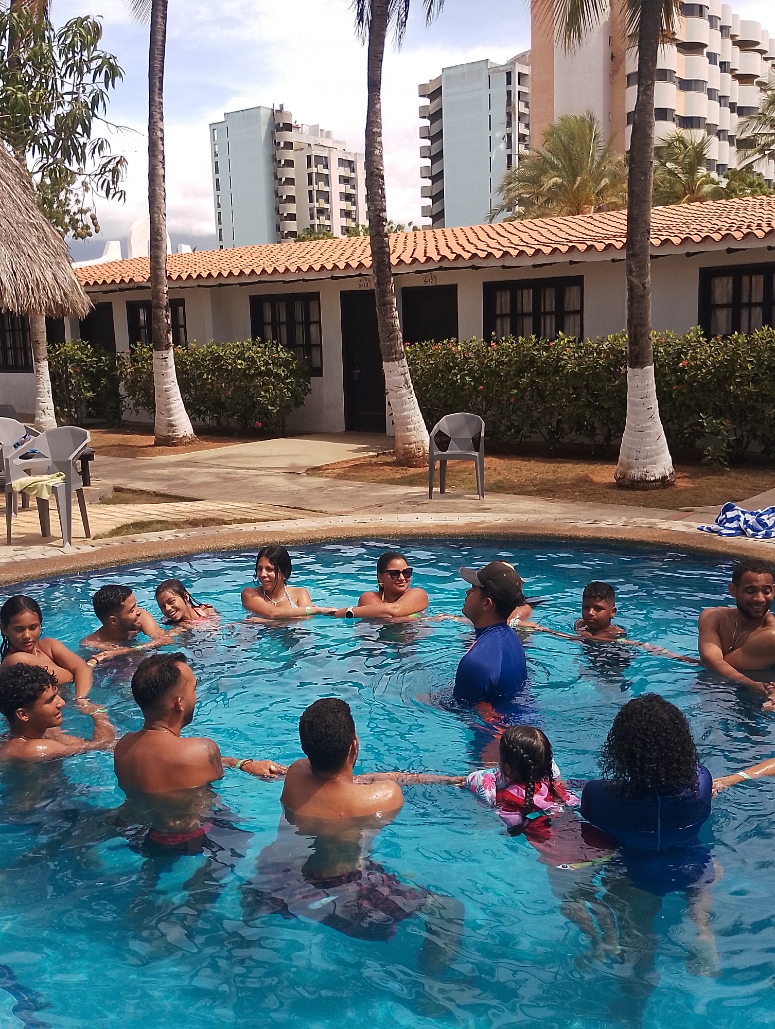 The image depicts an outdoor scene in Porlamar, Venezuela, centered around a swimming pool. Approximately 15 individuals, primarily adults with at least two children, are gathered in the clear blue water of a tiled swimming pool. Many are smiling, and several are holding hands, indicating a group activity. Attire consists of various swimsuits and rash guards.
Around the pool, a light-colored paved deck is visible, with green grass and manicured bushes beyond it. Two grey plastic chairs are positioned on the deck on the left side, and a blue and white striped towel rests on the deck on the right. Several bungalows or cabins with white walls and reddish-orange tiled roofs are located behind the bushes. These structures feature dark-framed windows and doors, with some displaying room numbers such as "5-10" and "5-12."
Tall palm trees with white-painted trunks are interspersed throughout the scene, along with another leafy tree bearing fruit, possibly mangoes, on the left. In the distant background, several multi-story buildings are visible under a partly cloudy sky. These include a light blue/grey building with a stepped design, a taller building with a distinct curvilinear balcony architecture, and other high-rise structures typical of an urban coastal area. The scene is brightly lit, suggesting daytime.