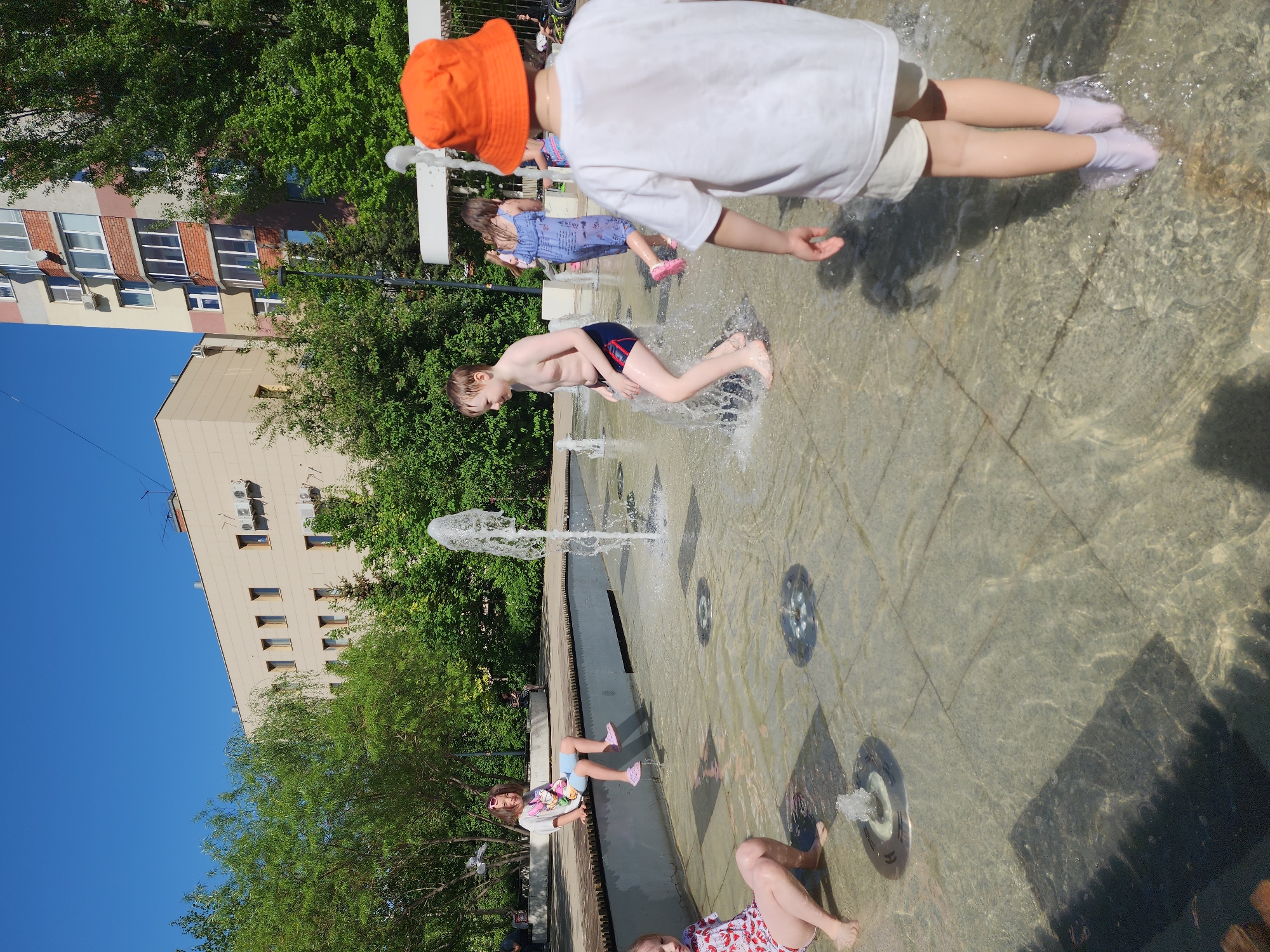 Here is a description of the image:
The image shows several children playing in a water fountain in an urban setting. The fountain appears to be located in a public square with buildings and trees visible in the background.
Several children are interacting with the water, some running through the fountain, some sitting at its edge, and at least one lying in the water. They are wearing various clothing, including swimwear, t-shirts, shorts, and socks, and one child has a bright orange hat.
The fountain is a modern design with multiple jets of water spraying upwards. The surrounding area is paved with stone or concrete tiles. The day is bright and sunny, casting shadows on the paved surface. The buildings in the background are multi-storied and appear to be residential or commercial. There are trees scattered throughout the scene, providing shade and adding to the park-like atmosphere.