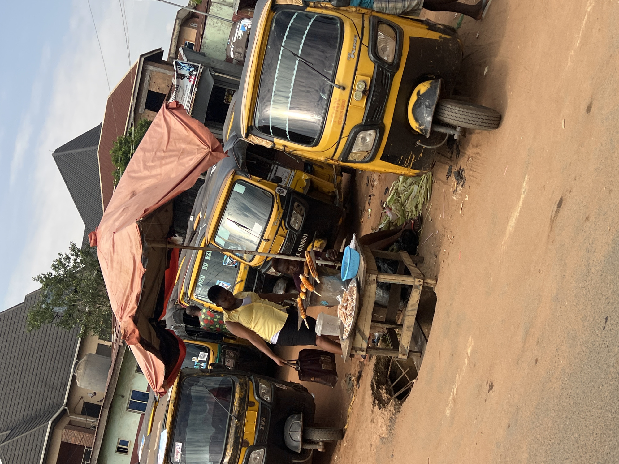 In Nkpor, Nigeria, a street scene depicts a collection of yellow auto-rickshaws, commonly known as "Keke Napep" or "Marwa," parked along a dusty road. The vehicles, displaying various registration numbers like "C. 408601" and "LAKU BY EZI," are positioned close together, suggesting a busy transport hub.
A woman in a yellow sleeveless top and black skirt stands beside a stall, holding a dark brown bag. The stall, constructed from wooden planks, displays a platter of what appears to be fried fish or meat, alongside several ears of corn roasting on sticks. Another individual, dressed in a purple shirt, is seated behind the stall, seemingly attending to the food.
The ground is a mixture of reddish-brown dirt and patches of sand, with a visible hole in the ground near the stall, possibly a drainage or manhole. A large, faded orange umbrella is propped up, providing shade over the street vendors and some of the parked Keke Napeps. In the background, multi-story buildings with distinctive roofing styles are visible, hinting at the urban environment. The sky is a pale blue with scattered clouds.