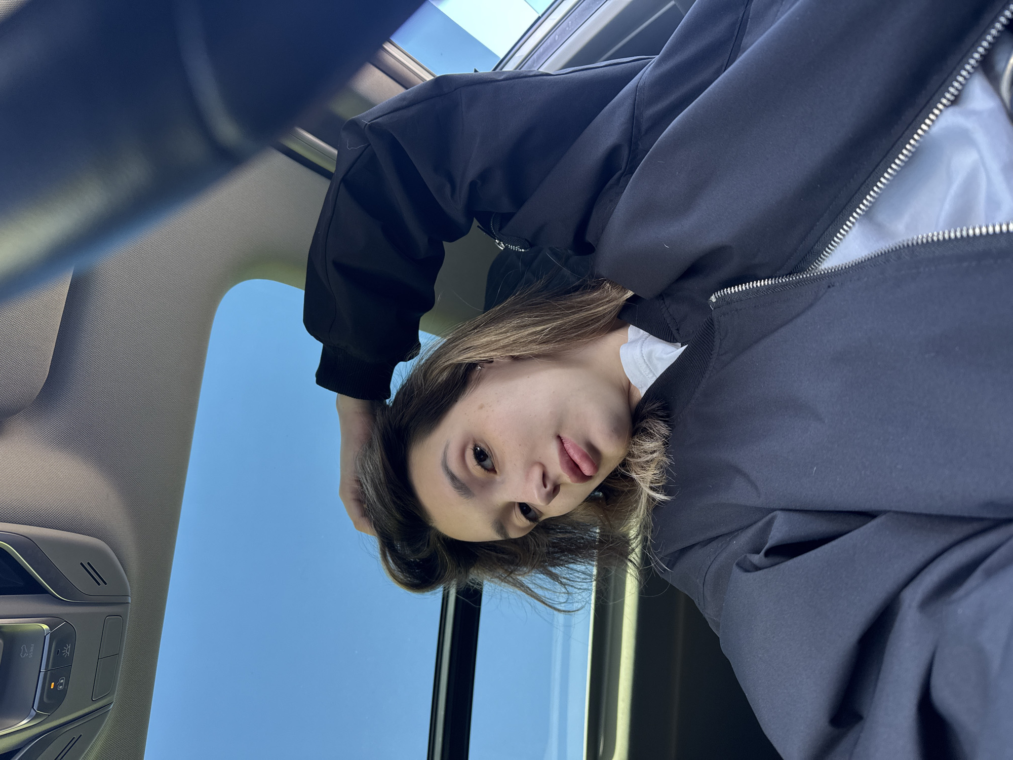 A young person, likely female, is featured in a playful portrait taken from an upward angle inside a vehicle. The subject, with light brown hair, looks directly at the camera with a slight head tilt and a playful expression, her tongue peeking out slightly. She is wearing a black bomber-style jacket with a visible silver zipper over a white undershirt.
The setting is the interior of a car, specifically looking up towards a panoramic sunroof. Through the sunroof, a clear, bright blue sky is visible, indicating daytime and good weather conditions. Parts of the car's interior are seen, including a grey textured headliner and an overhead control panel. This panel features modern buttons, including one with a lightbulb icon, suggesting controls for interior lighting, and another resembling an emergency call button or assistance service, along with small speaker grilles. The lighting in the image is bright and natural, coming from the open sky above. The subject's hand is visible behind her head, possibly holding her hair or simply part of the pose. No discernible text is visible in the image.