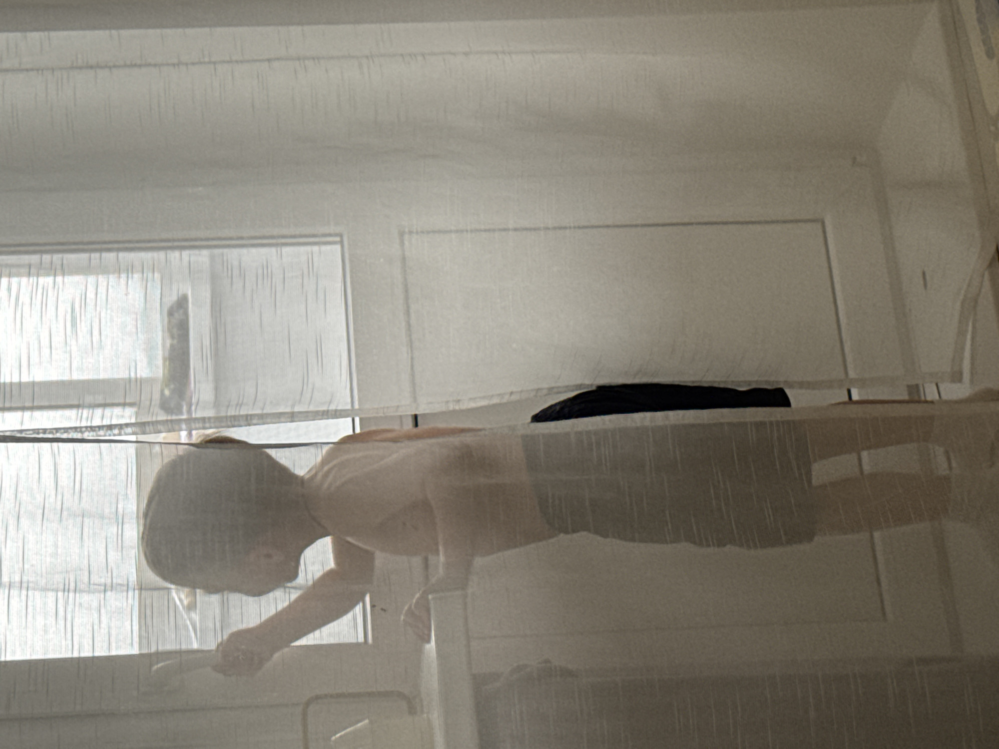 A child is standing in front of a window, partially obscured by a sheer curtain. The child is wearing dark shorts. The room appears to be an interior space with a door visible in the background. The location is Chaikovsky, Russia.