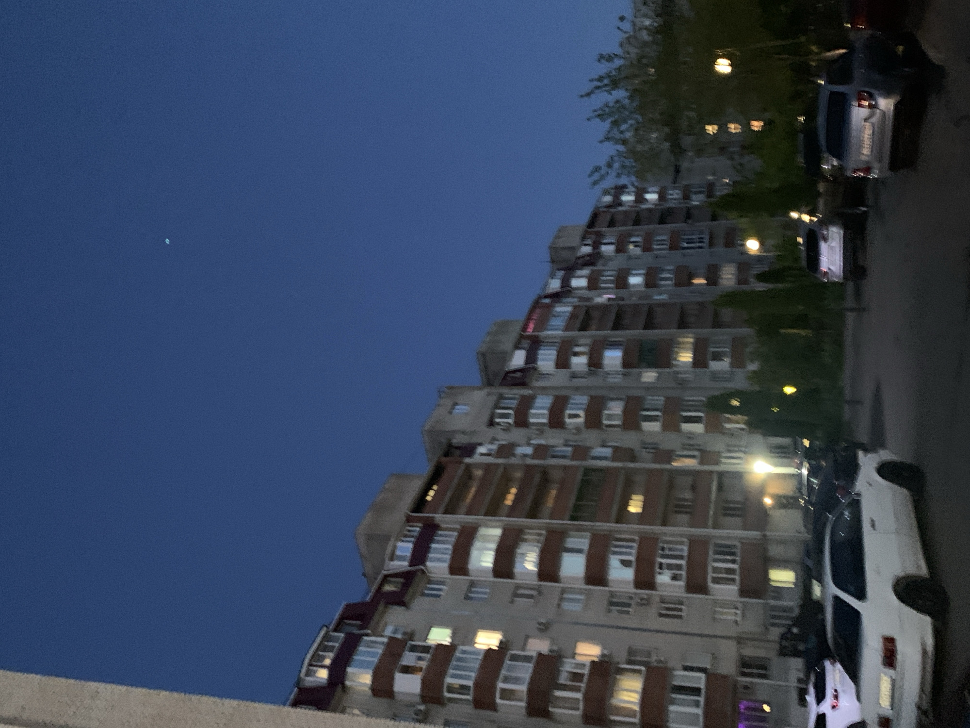 The image, rotated 90 degrees counter-clockwise, presents an urban residential scene at night or late dusk in Stavropol, Russia. A large, multi-story apartment building dominates the left and central parts of the frame. Its architecture features a light grey concrete facade with distinctive dark red/brown horizontal bands or balconies, and a staggered or stepped design. Many of the building's numerous windows are illuminated with warm light, indicating occupied units.
Adjacent to the building, on what appears to be a street or parking lot, several cars are parked. A white sedan is prominent in the right foreground, with its rear visible. Its license plate shows partially discernible characters, including "016" or "O16" at the beginning. Behind it, a darker car is also parked. Scattered artificial lights, likely streetlights or exterior building lights, cast soft yellow and orange glows in the area around the vehicles and trees.
The sky above is a deep, clear indigo blue, signaling a clear evening or night. A small, blurry, light-blue speck, possibly a distant star, planet, or an atmospheric light, is visible in the upper left section of the original image. Trees with dark green foliage are present, intermingled between the building and the parked cars. The scene is static, depicting a tranquil moment in a residential area with no explicit activity or event taking place.