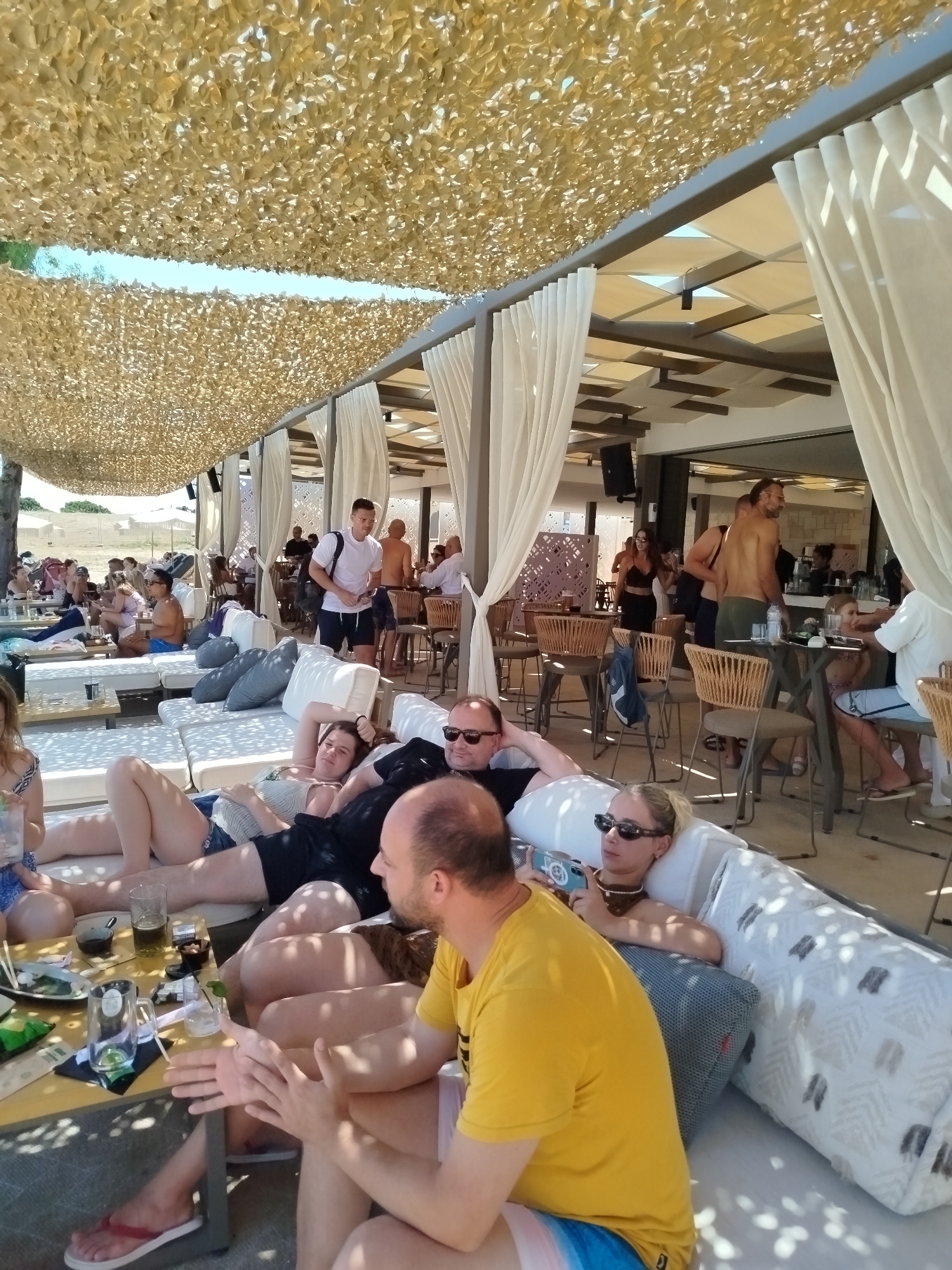 In Sithonia Municipal Unit, Greece, a group of people are gathered at an outdoor cafe or beach club. The scene is shaded by a textured canopy overhead. In the foreground, three individuals are seated on a white sectional sofa. A man in a yellow shirt faces left, with his hands clasped. To his right, a woman with blonde hair and sunglasses is reclined on the sofa, looking at her phone. Another person is partially visible to the far right. In the middle ground, a man in a black shirt and shorts is also reclining on the sofa, with another person next to him wearing a black top and denim shorts. Further back, several other individuals are seated at tables or standing. One man in a white t-shirt and dark shorts stands holding a bag. The environment suggests a relaxed, sunny day at a coastal establishment.
