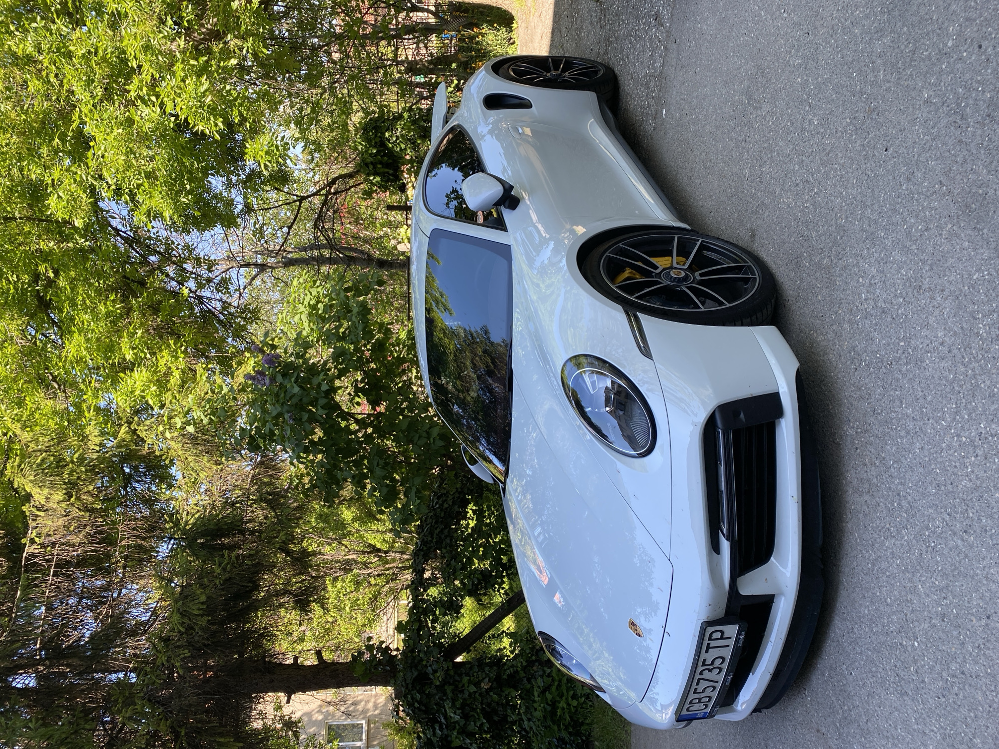 The image shows a white Porsche 911 GT3 parked on a paved road. The car is positioned at an angle, facing the camera, with its front end near the bottom of the frame. The license plate reads "CB 5735 TP." The background consists of lush green trees and vegetation, with glimpses of a building visible through the foliage on the left side of the frame. The image was taken outdoors on a sunny day, with some shadows cast on the car and the road. Given the license plate and the surrounding environment, the location is likely in a residential or suburban area in Pleven, Bulgaria.